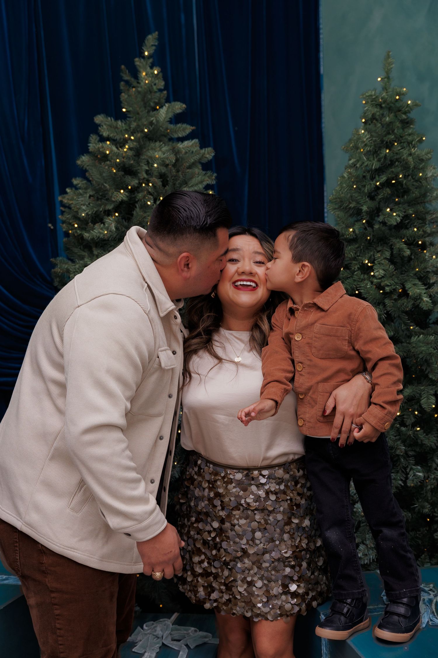 A family shares loving kisses and embraces in front of decorated Christmas trees in a holiday photo session.