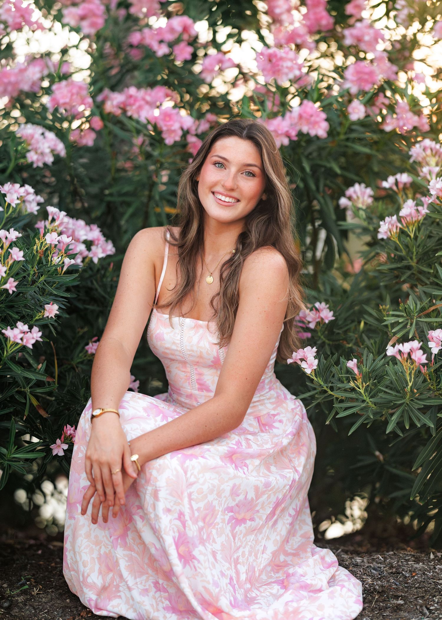 Smiling woman with long brown hair in a pink floral sundress sits among blooming pink oleander bushes in soft golden sunlight.