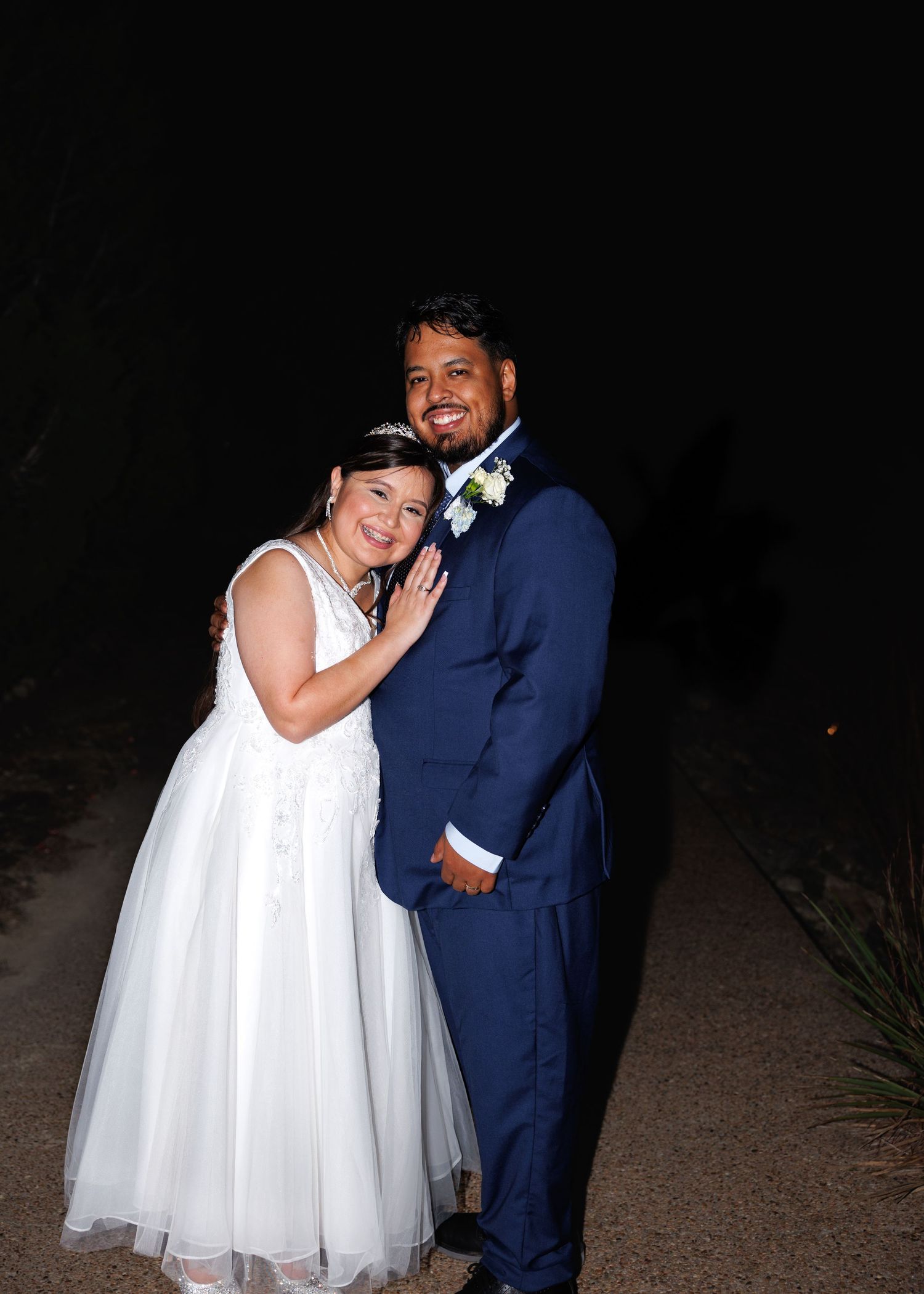 A couple shares a joyful moment in evening attire, with the bride in white and groom in navy blue against a dark background.