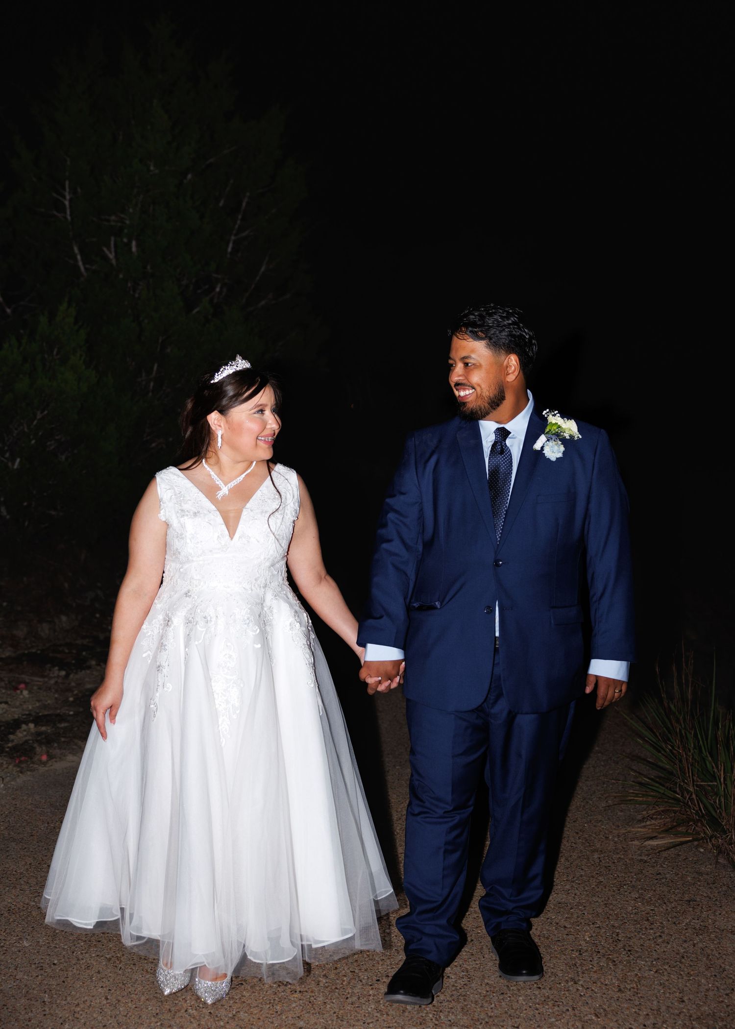 A wedding couple walks along a dimly lit outdoor path while holding hands wearing white dress and navy suit.