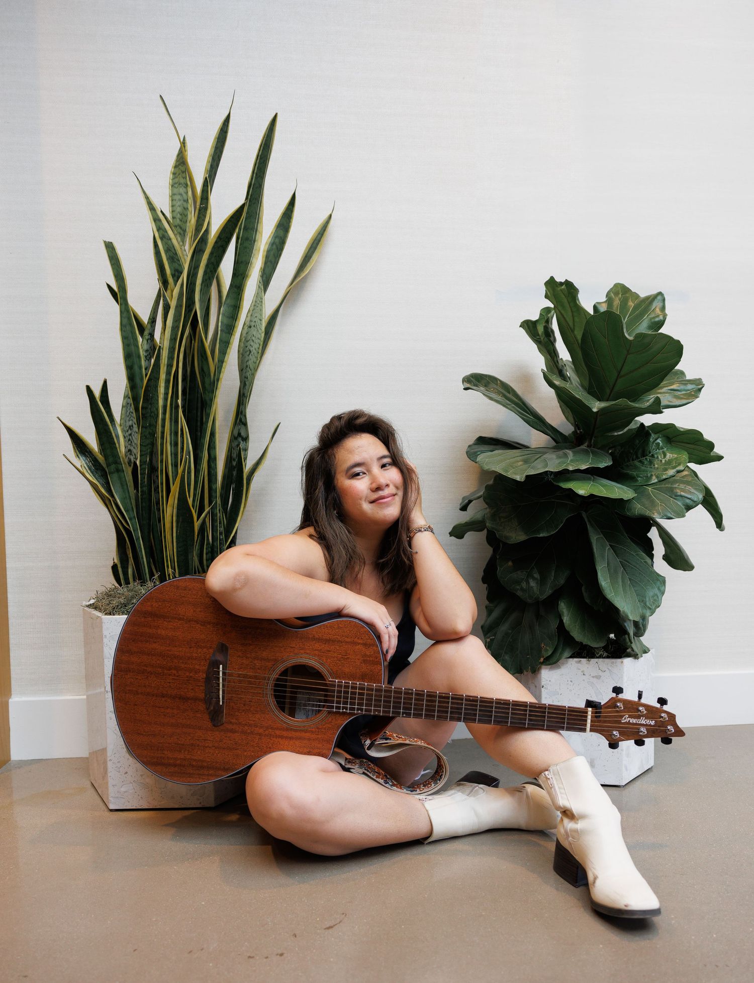 A person lounges on the floor with a wooden guitar and decorative indoor plants in a minimalist setting.