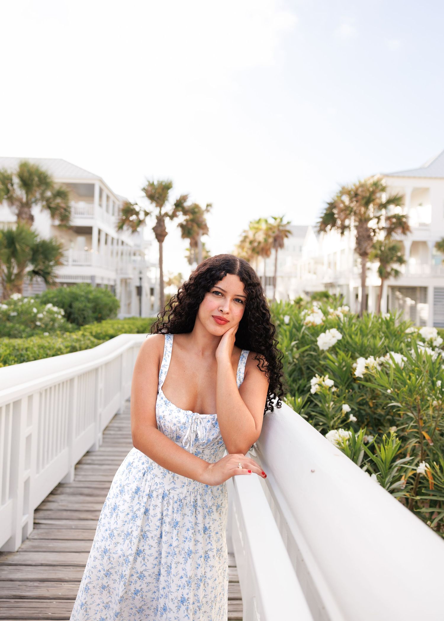 Person in a blue and white floral sundress poses on a coastal boardwalk with palm trees and seaside buildings in background.