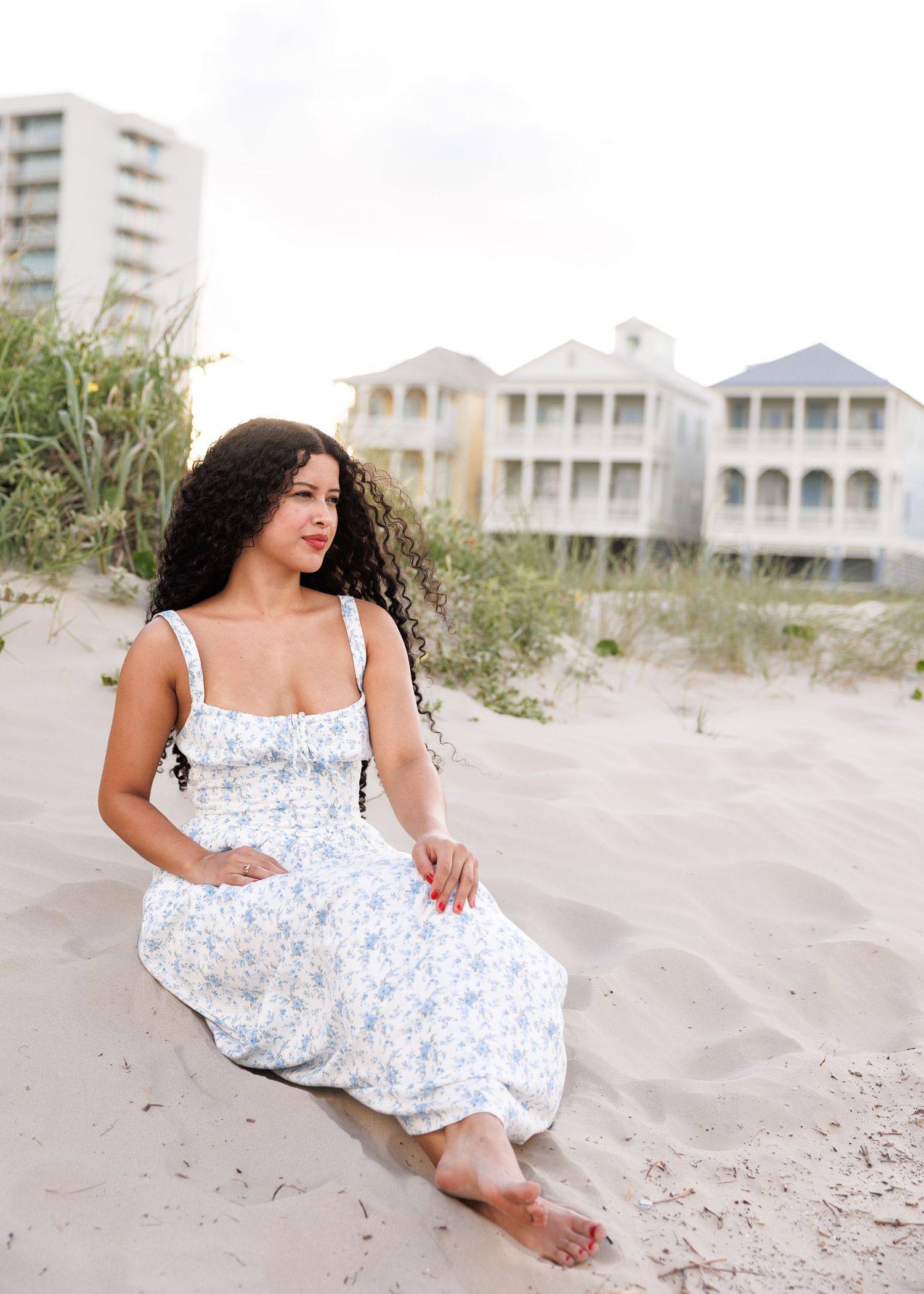 A woman in a white floral sundress sits gracefully on a sandy beach with coastal homes in the background.