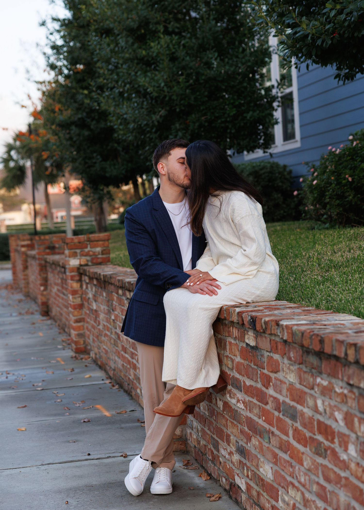 A couple sits together on a rustic brick wall in a romantic outdoor setting wearing navy and white outfits.