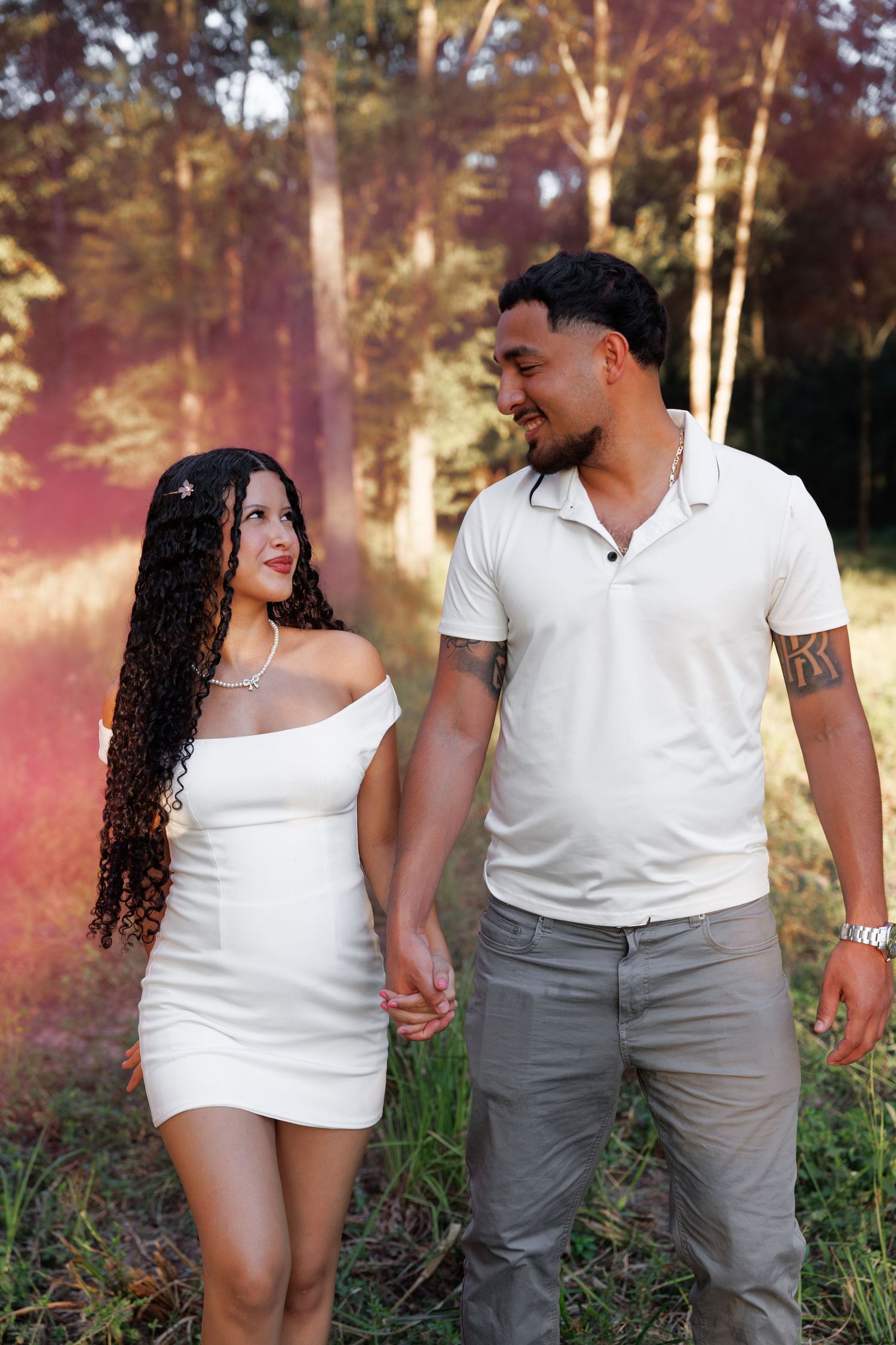 A couple in white outfits holds hands while walking through a sunlit wooded area on a romantic afternoon.