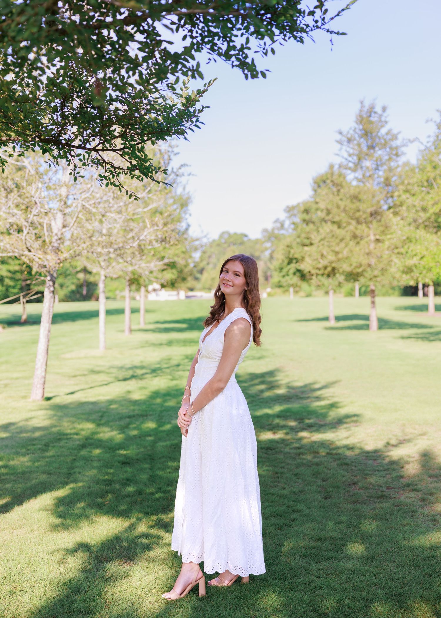 Woman in a long white sleeveless dress and tan heels stands smiling on a sunny grassy lawn surrounded by tall green trees.