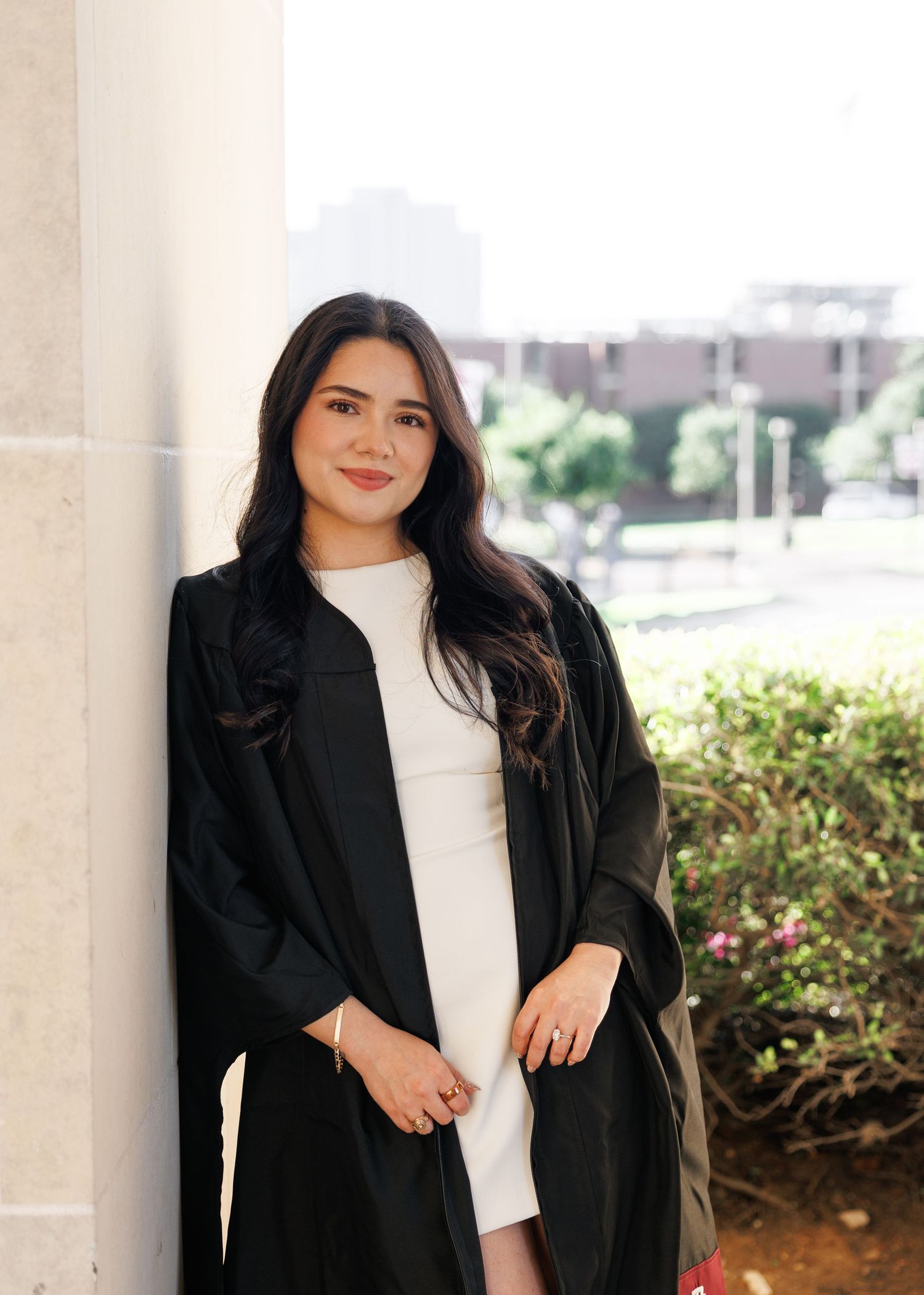 Young woman in graduation gown and white dress smiling outdoors near columns and greenery.