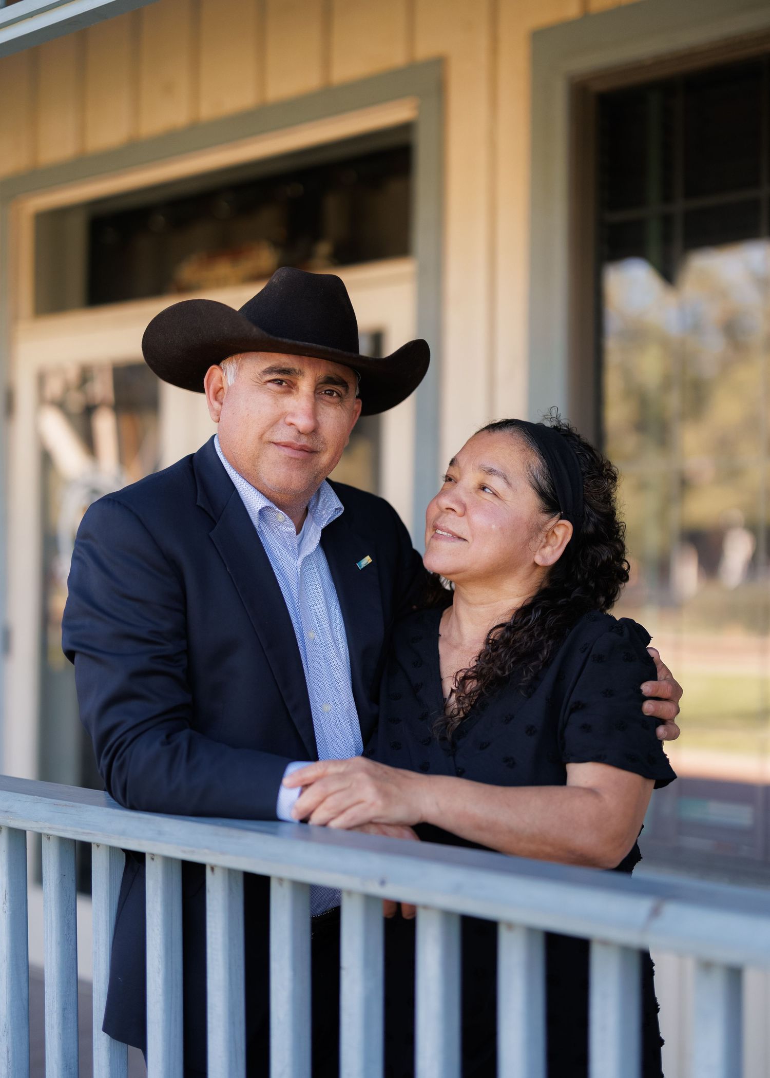 A couple in western attire lean together on a porch railing, sharing an intimate moment outside a rural building.