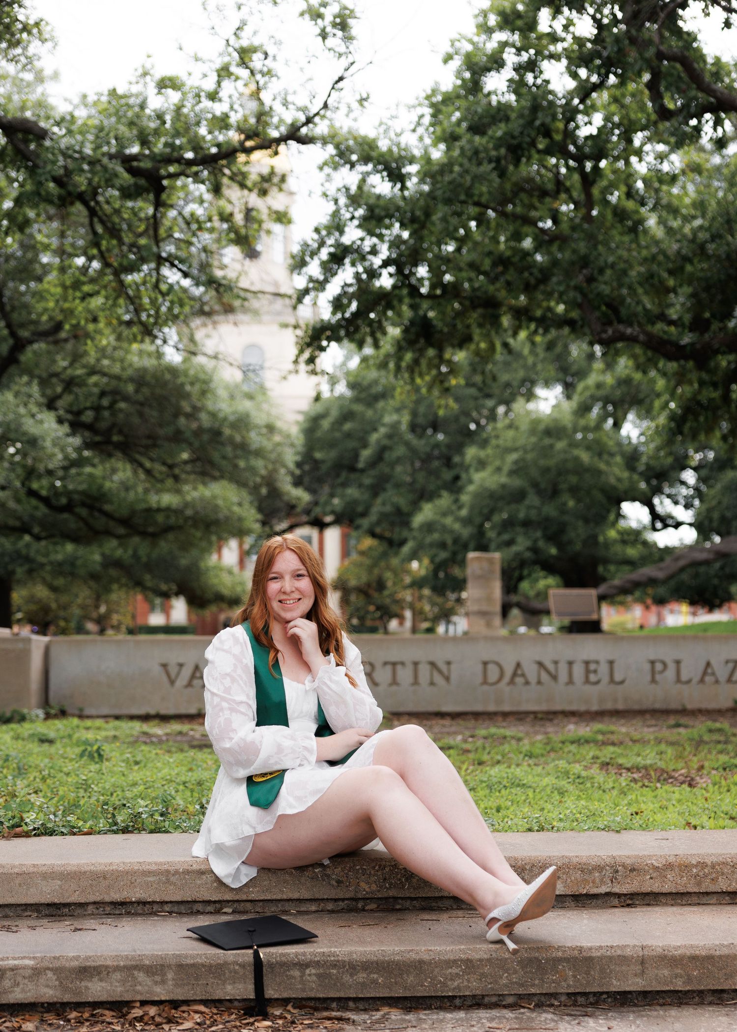 A person wearing a white blazer and green top sits on stone steps outdoors under overhanging trees at a plaza.