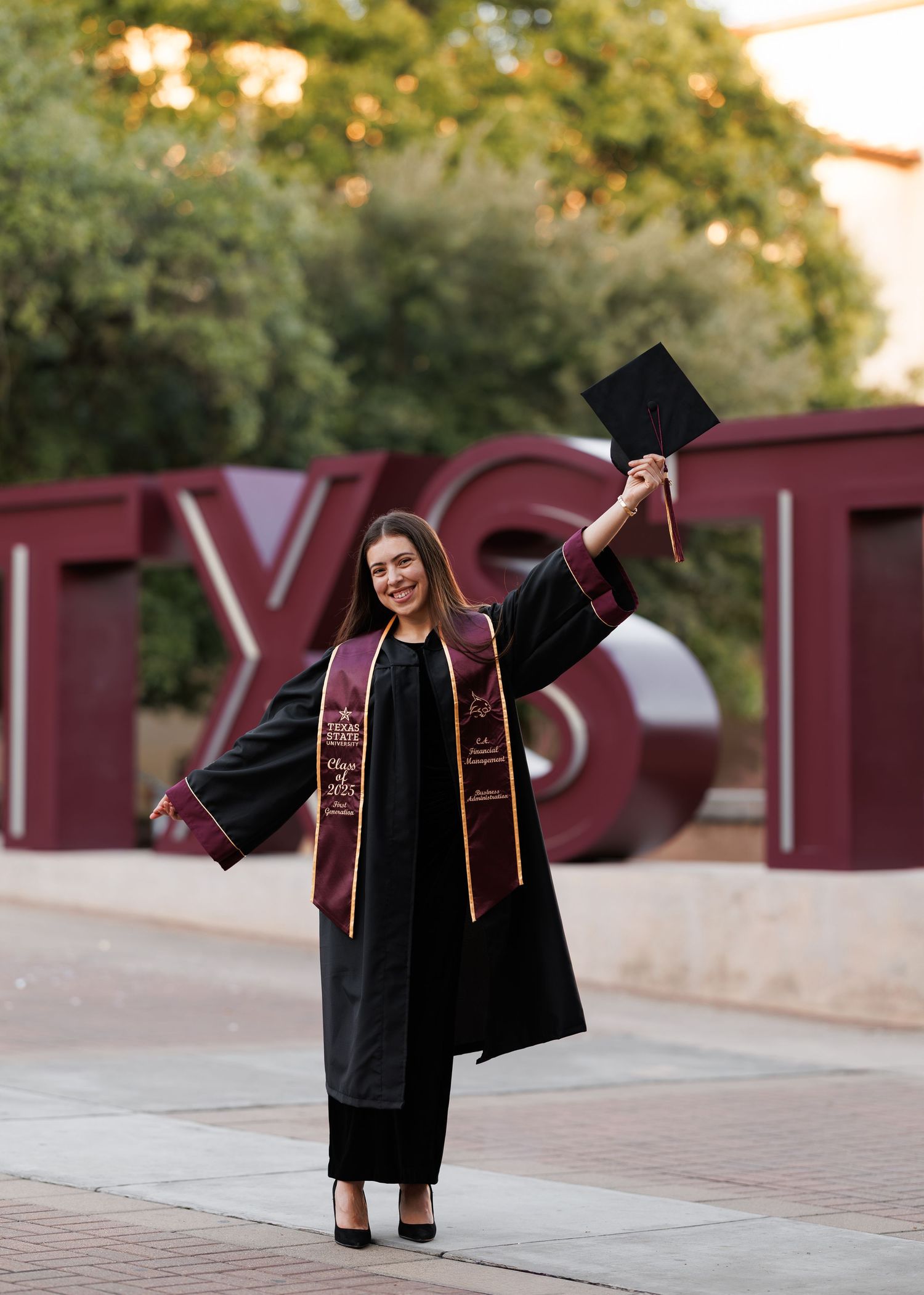 Graduate in black cap and gown poses in front of maroon university sign during outdoor graduation photoshoot.