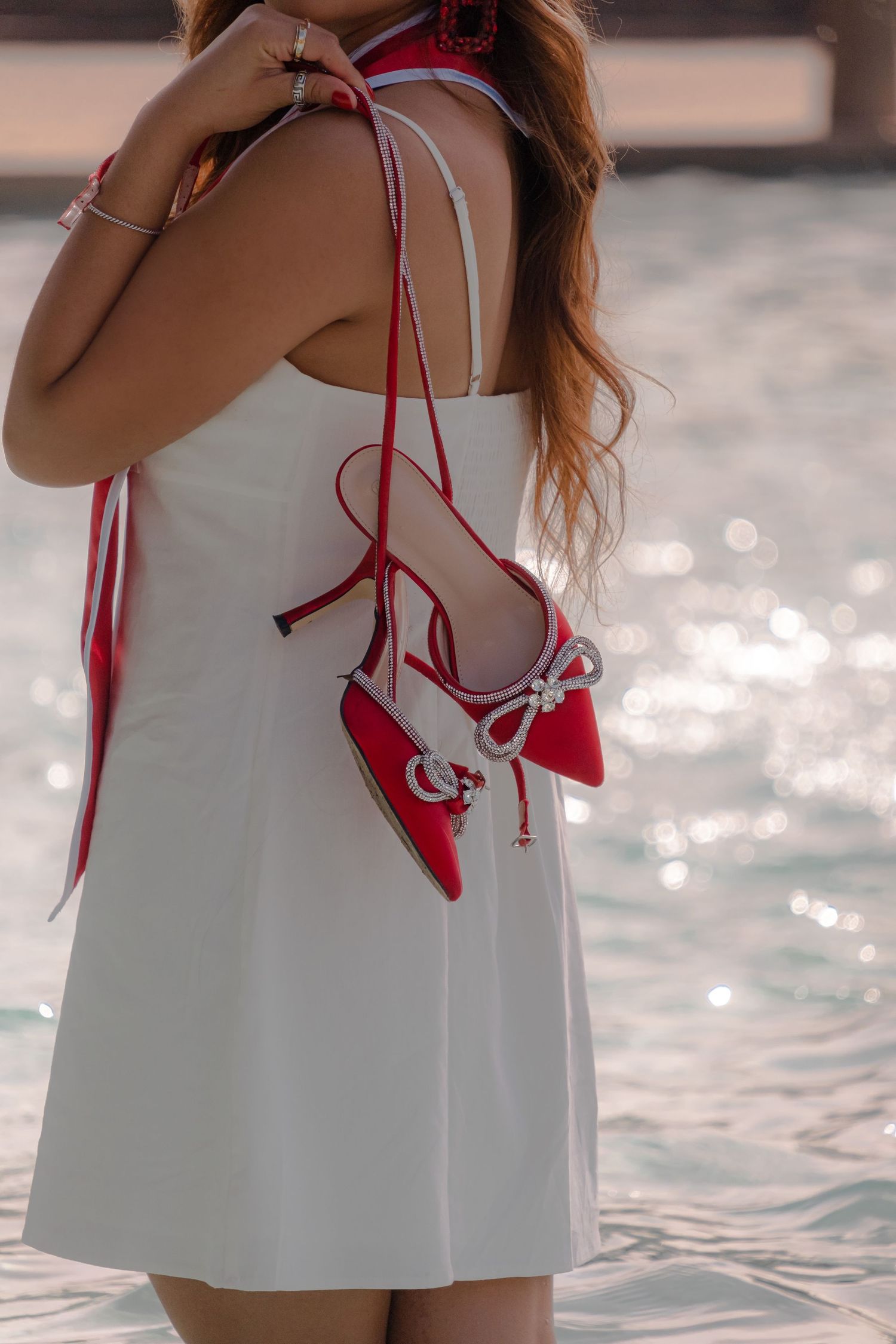 Woman in white summer dress holds red canvas sneakers with laces by sparkling water in sunlight.