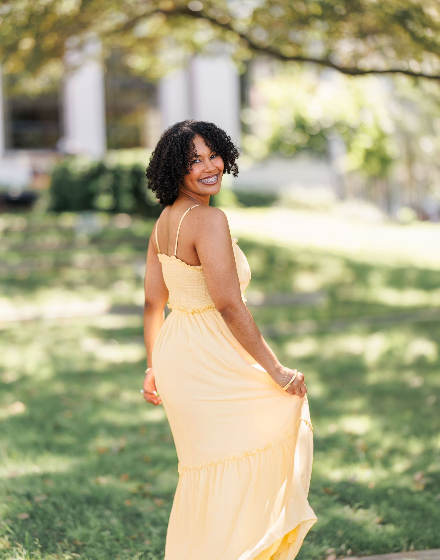 Young woman in a yellow maxi dress smiling outdoors in a lush green park setting.