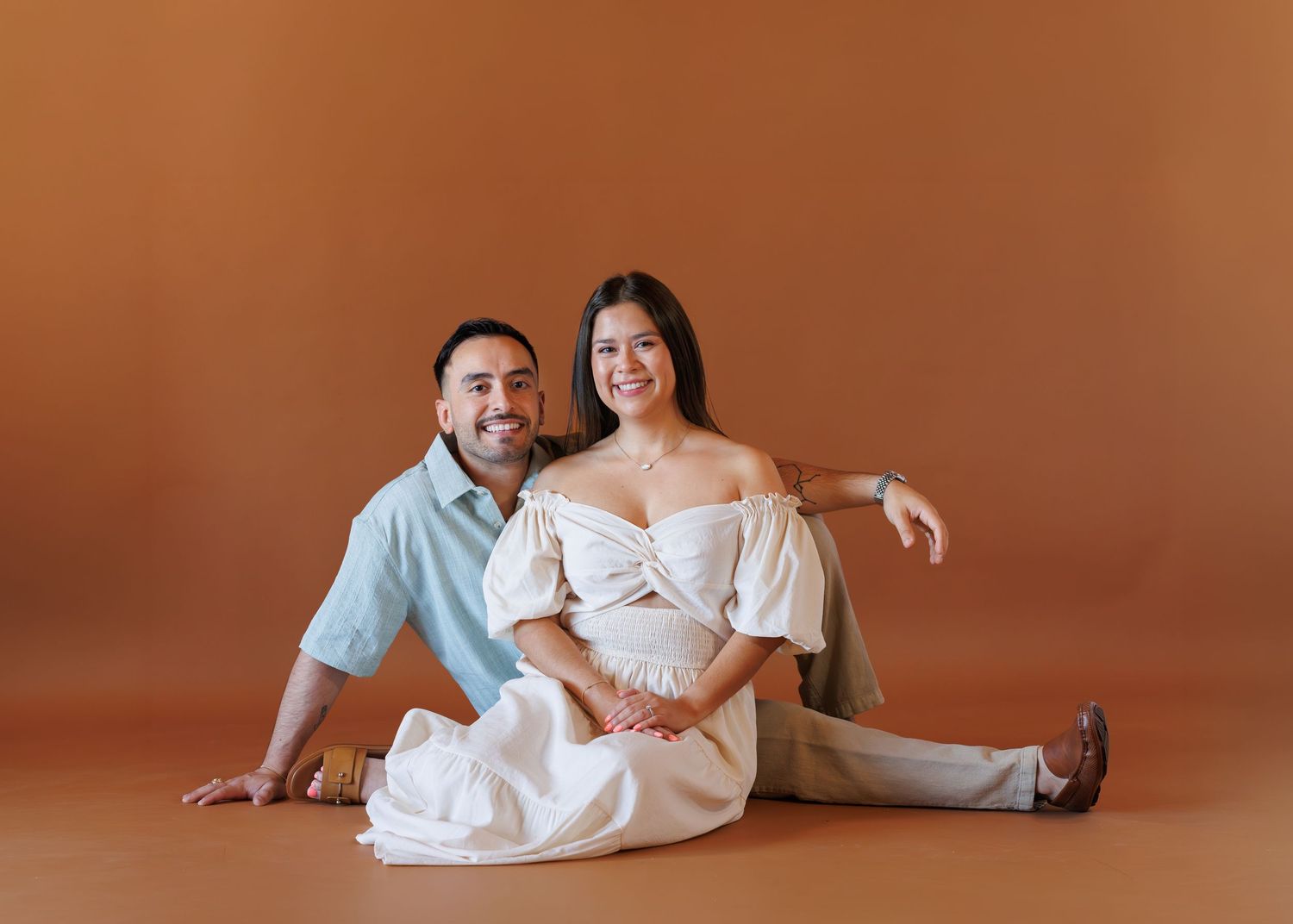Smiling couple posing for maternity photo shoot on warm brown background, woman in white off-shoulder dress cradling baby bump.