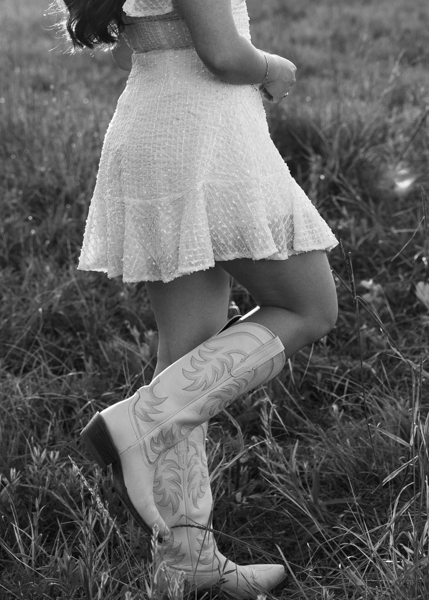 Black and white photo of someone wearing a white lace dress and decorative cowboy boots standing in a grassy field.