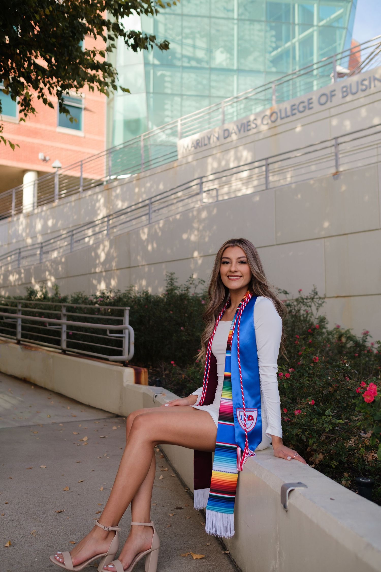 Graduate in white dress and colorful graduation stole sits on concrete ledge outside modern university building.