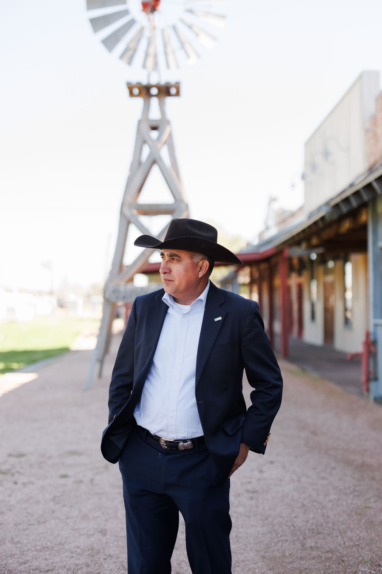 A cowboy in a dark suit and hat stands on a dirt path in front of a rustic storefront with a windmill in the background.