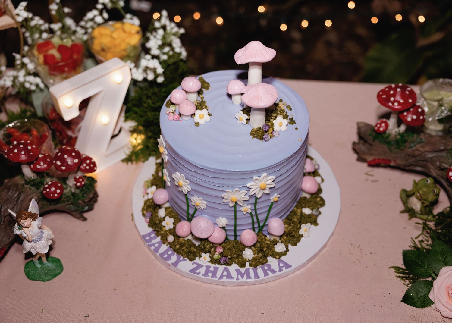 Purple mushroom-themed birthday cake with daisies and pink toadstools for Baby Zhamira, displayed at a fairy garden party.