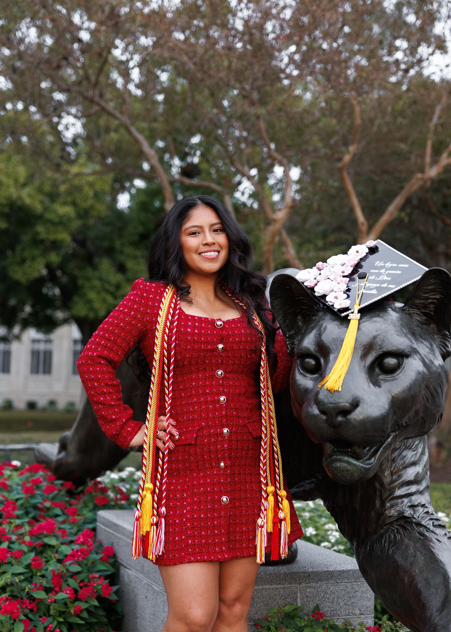A woman in a red crocheted top poses with braided hair near a black panther statue and rose garden on a college campus.