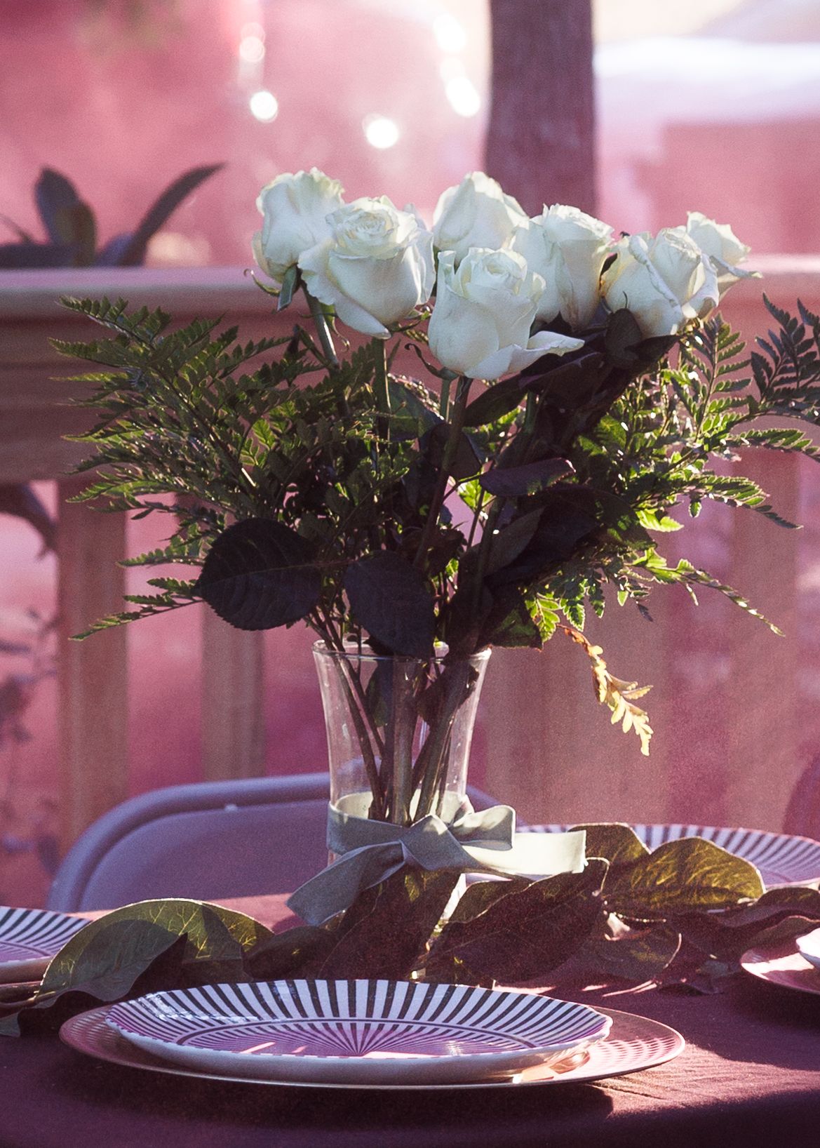White roses and ferns in a glass vase on a table set with striped plates in pink ambient lighting.