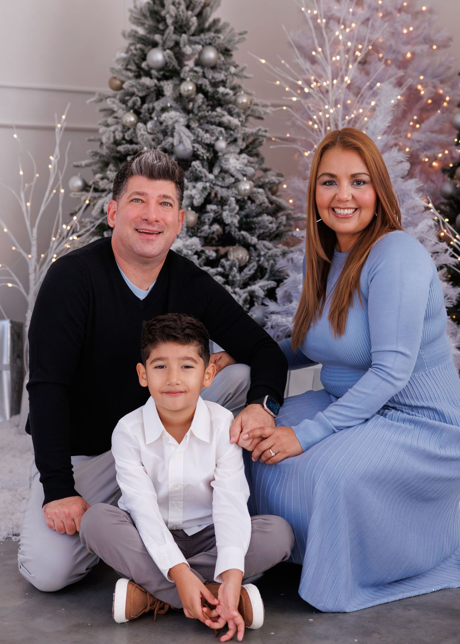 A family poses together in front of snow-covered Christmas trees and twinkling lights in a holiday photo session.