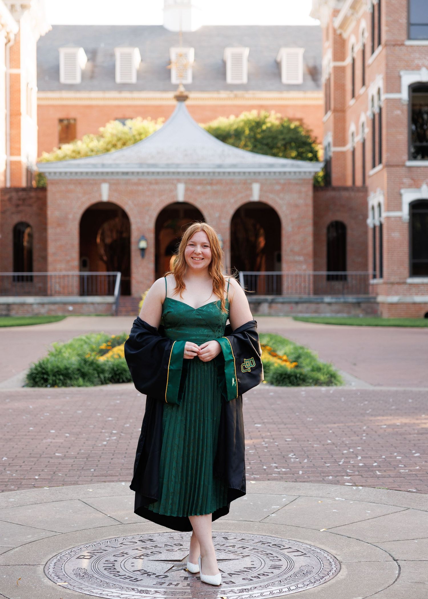 Graduate wearing black academic regalia and green pleated dress poses in front of brick university building with gazebo.