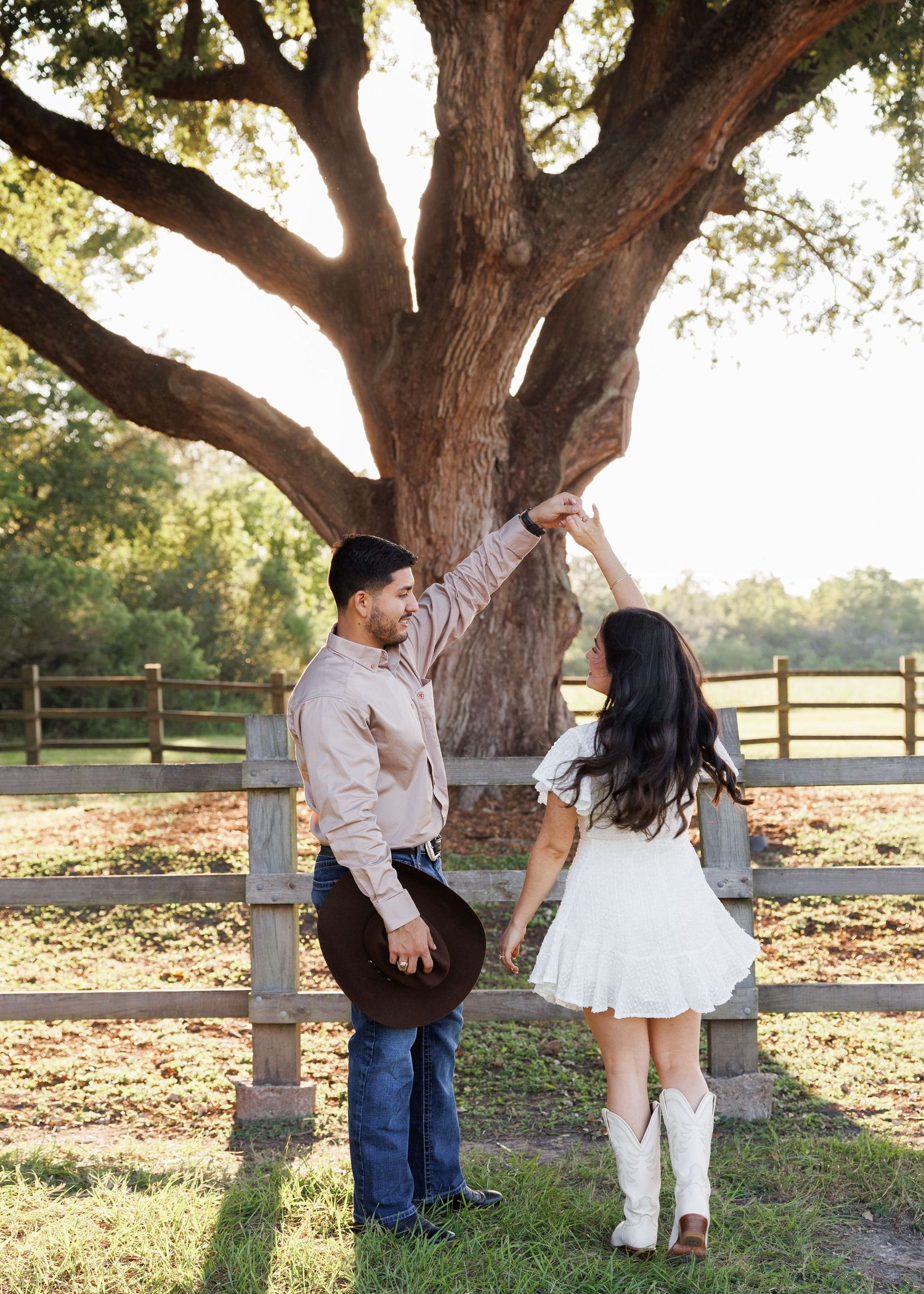 A couple shares a romantic moment under a large oak tree at sunset with wooden fencing in the background.