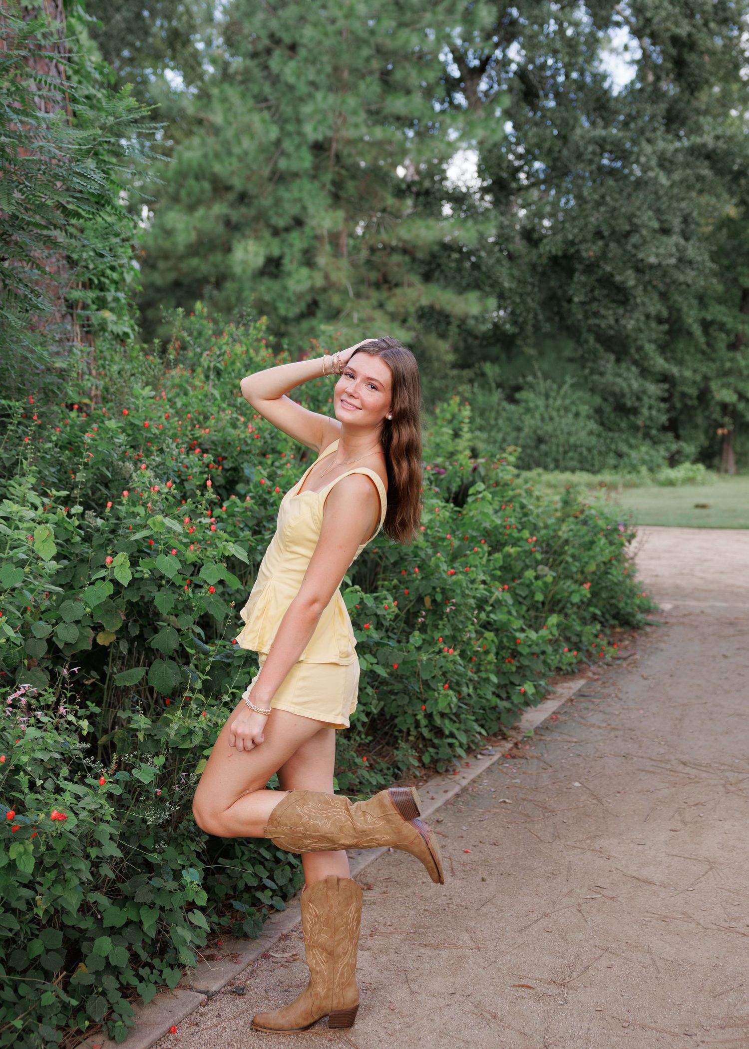 Young woman in a yellow romper and tan cowboy boots poses with one hand in her hair on a gravel garden path lined with green shrubs.