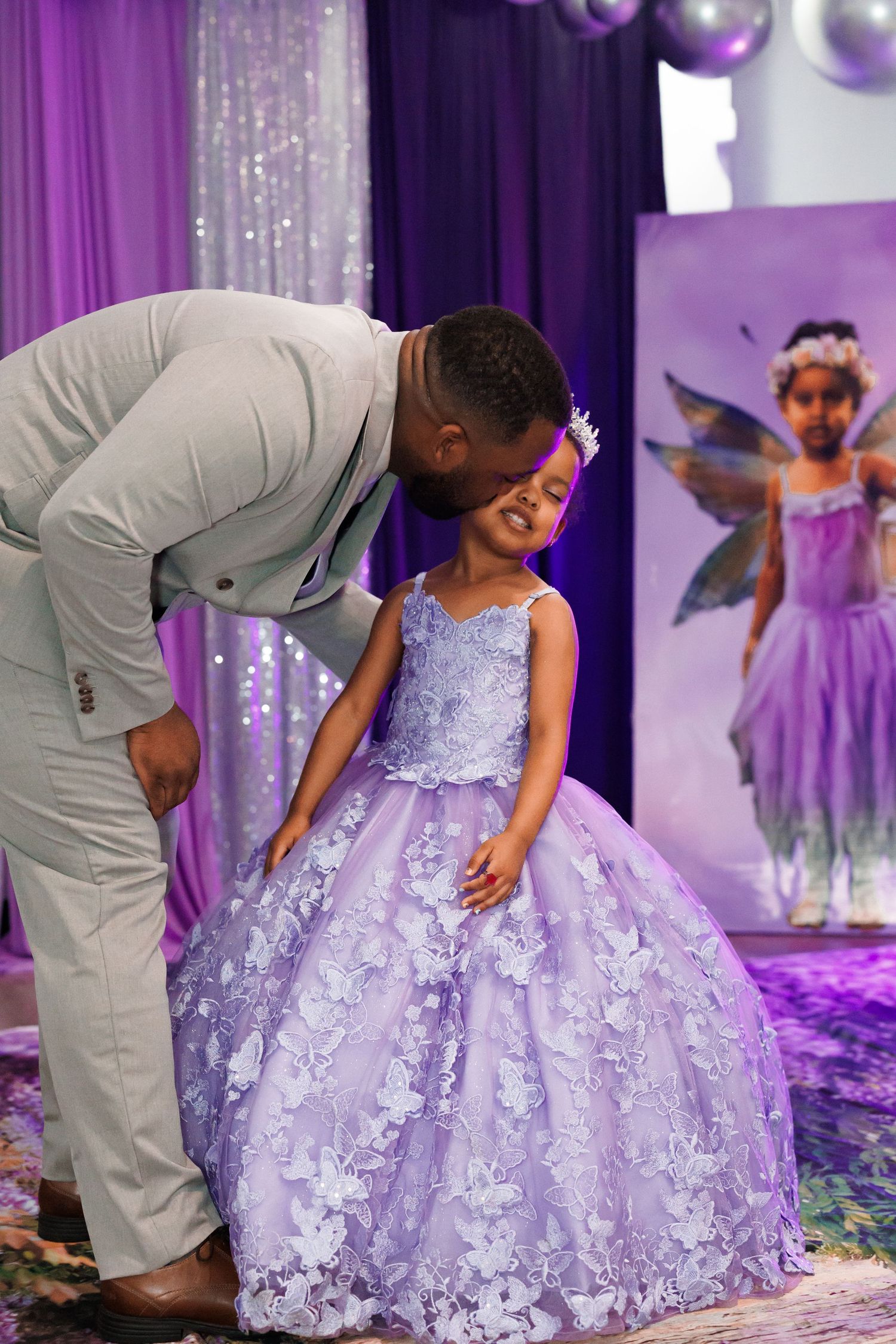 A fancy purple ballgown-style dress with floral lace details is worn at a fairy-themed celebration with sparkly decorations.