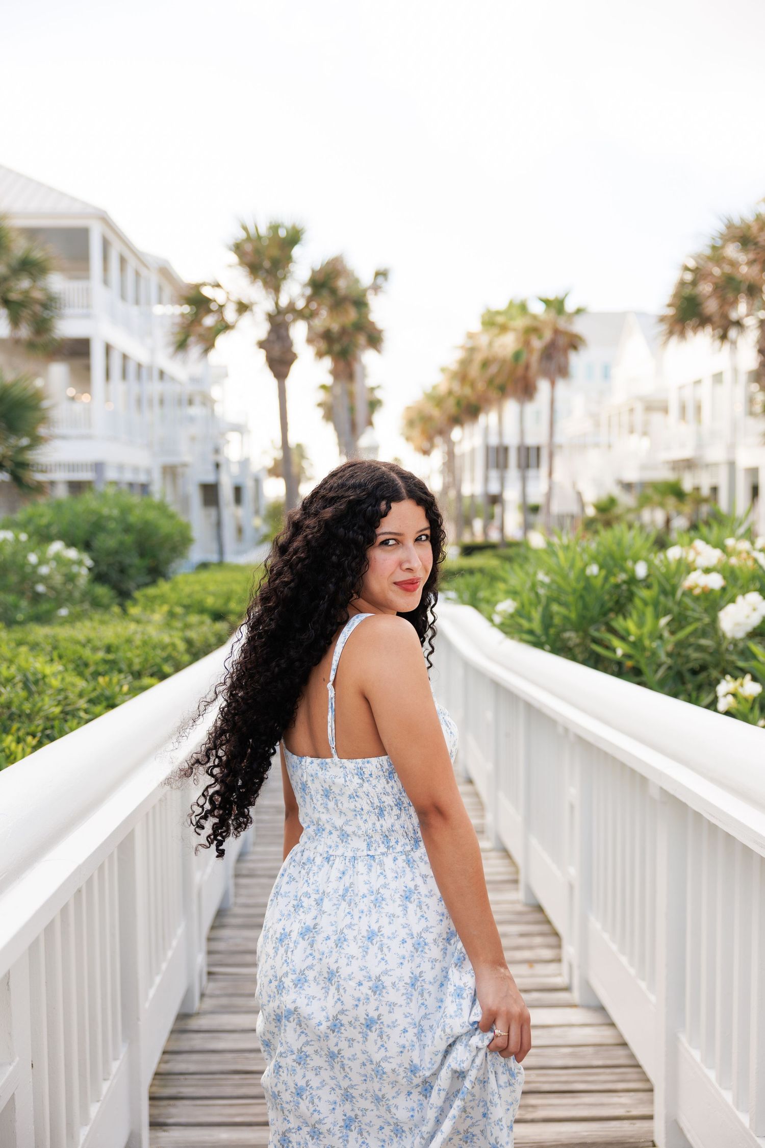 A model in a floral blue and white sundress poses on a white boardwalk with palm trees and beach houses in the background.