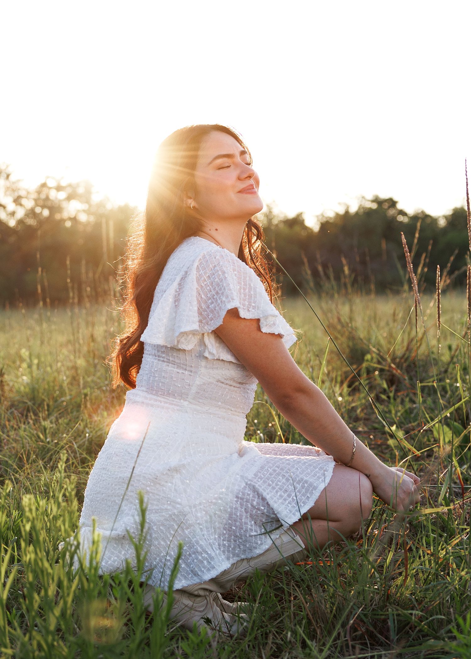 A person in a white dress sits in a sunlit field during golden hour.