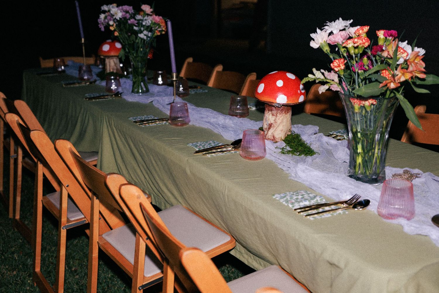Elegant long dining table with green tablecloth, mushroom decorations, floral centerpieces, and wooden chairs for an enchanted forest party.