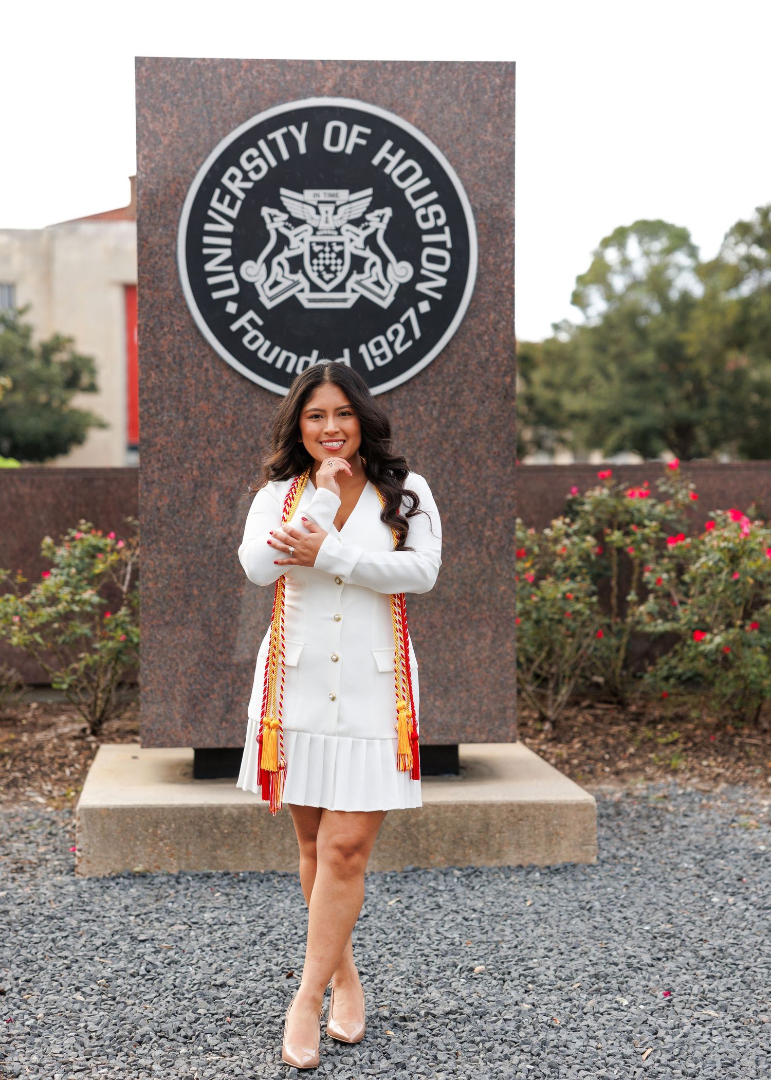 A graduate poses in white dress with accent sash in front of University of Houston seal and rose garden.