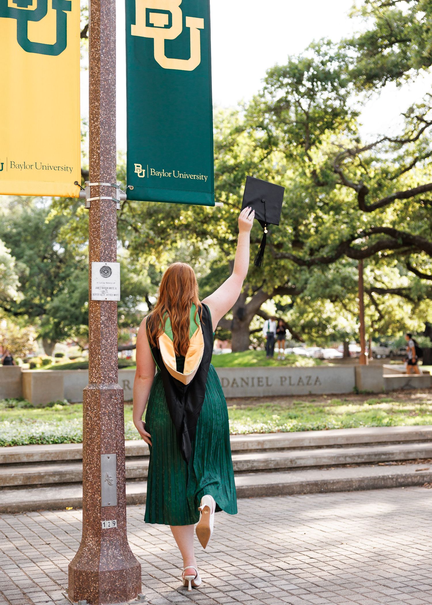 Graduate celebrating in green cap and gown poses near Baylor University campus banners on a sunny day at Daniel Plaza.