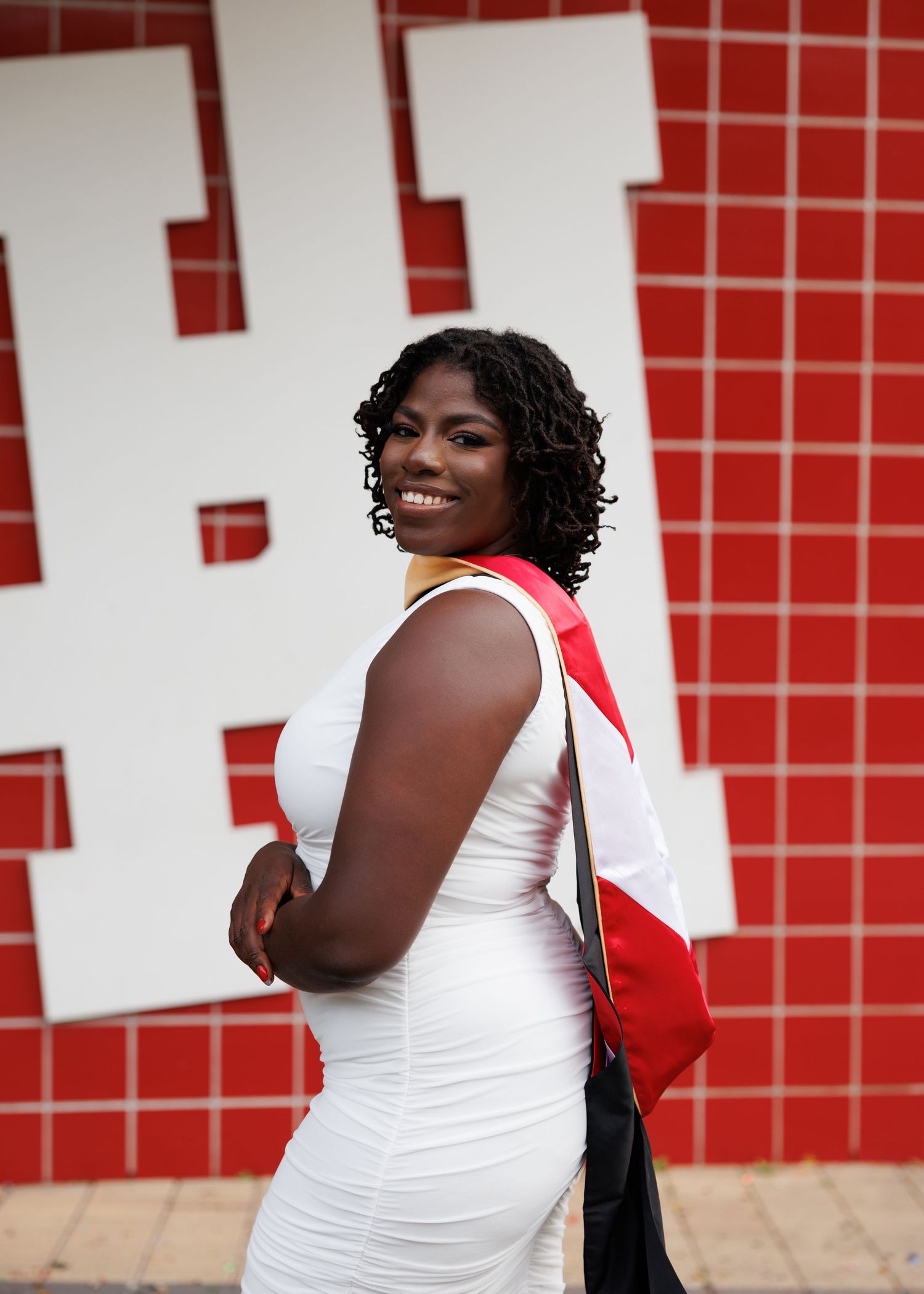 A person in a white dress poses confidently in front of a red tiled wall with a red shoulder bag.