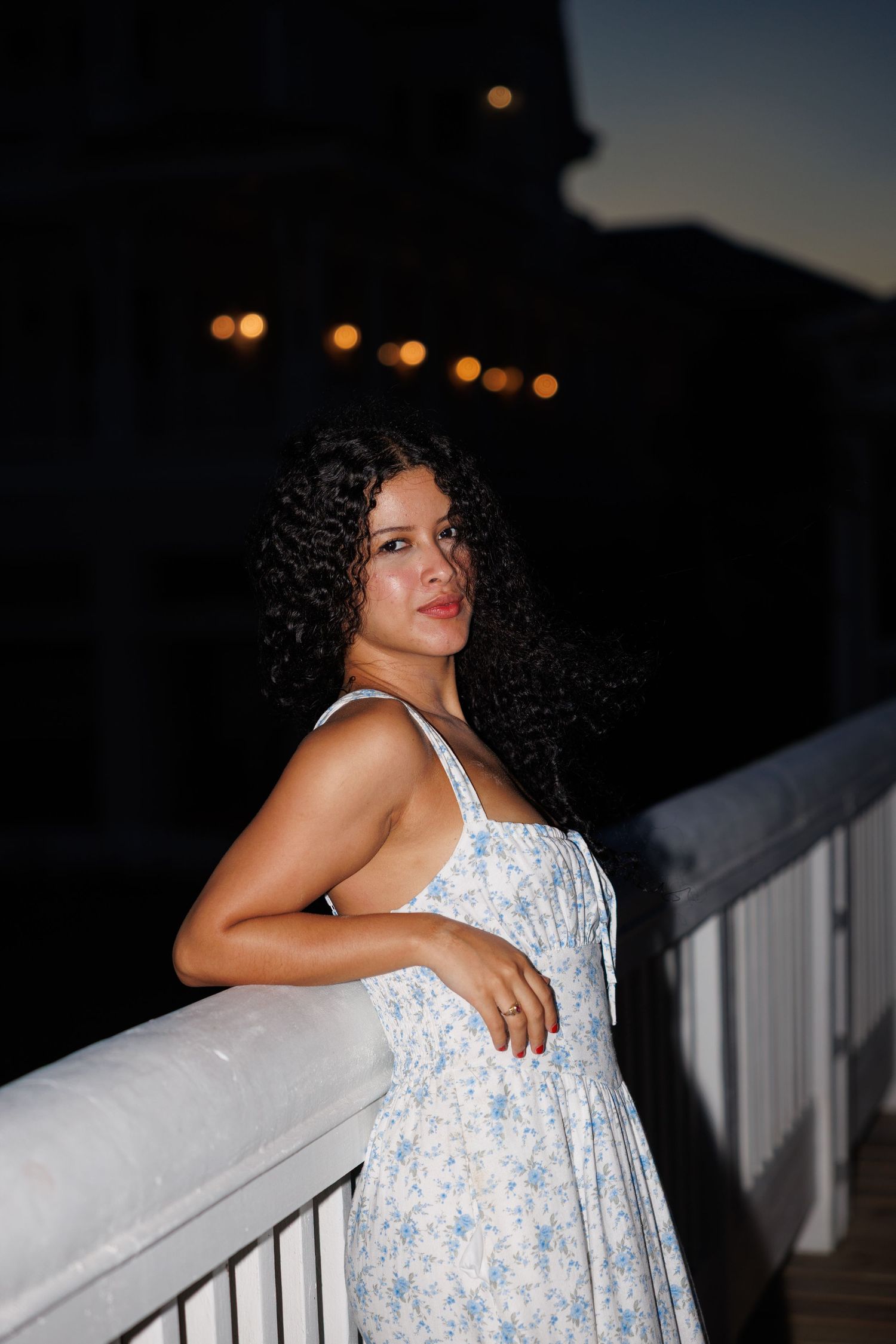 Person in a white floral sundress poses against a railing at dusk with glowing lights in the background.