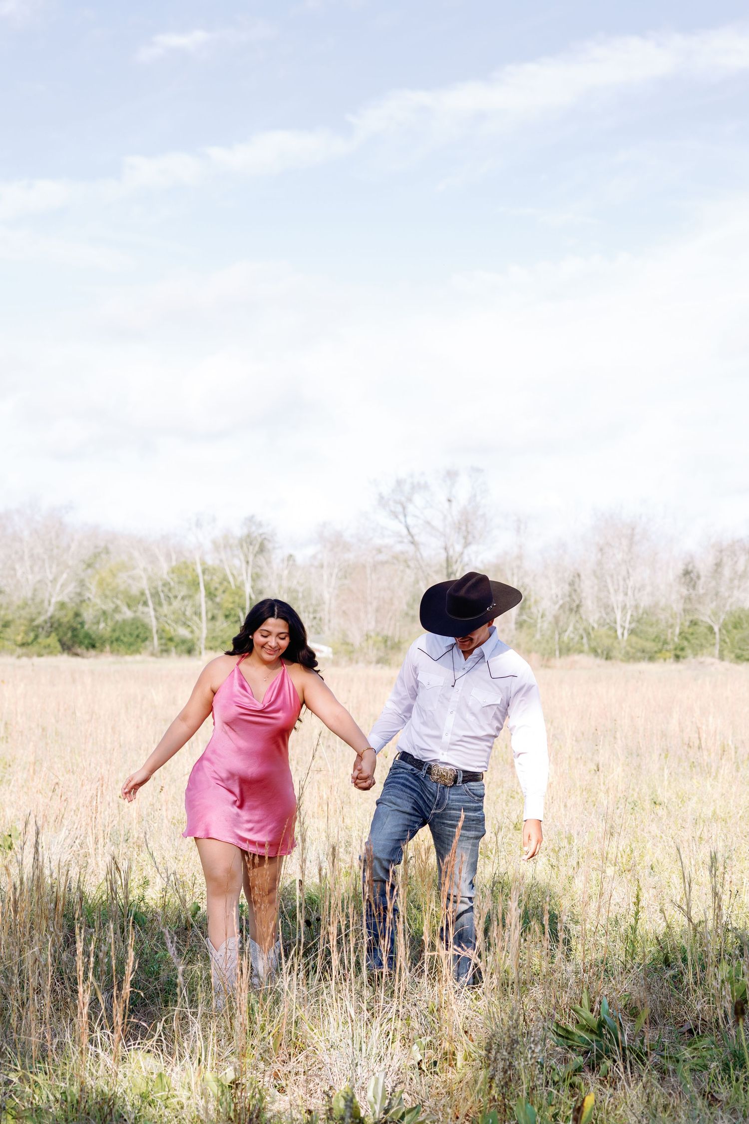 Couple holding hands walking through a golden field, woman in pink dress and man in cowboy hat and jeans.