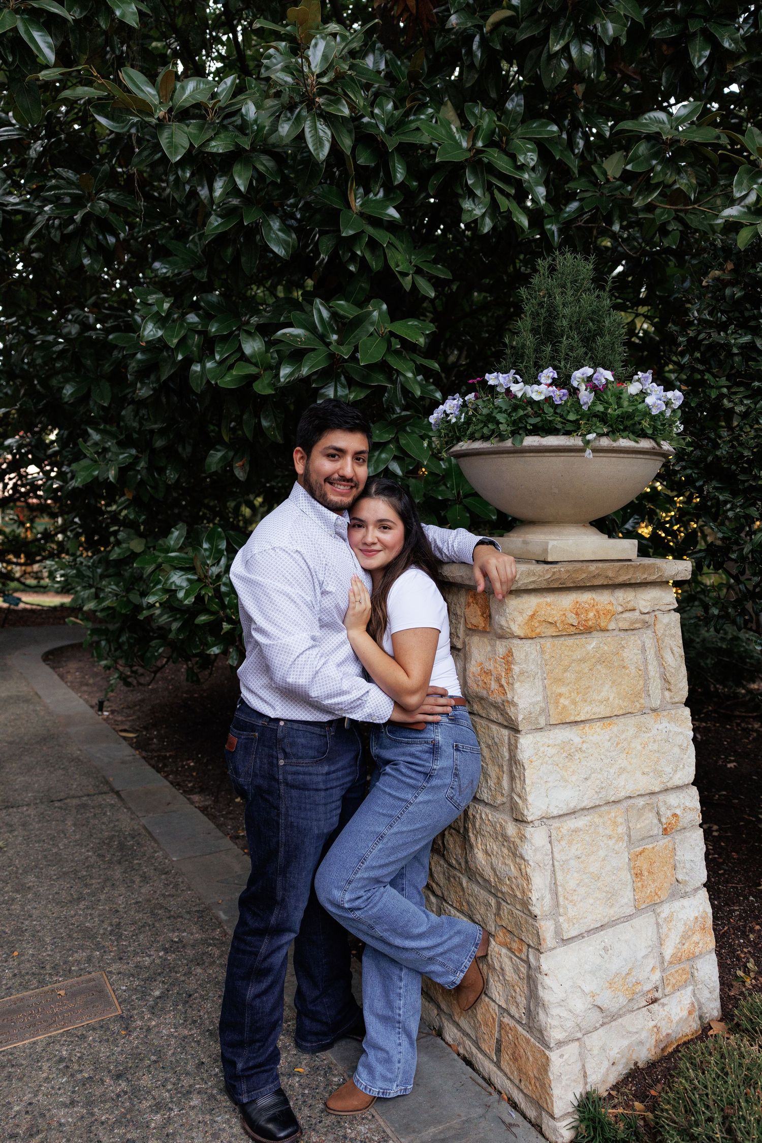 A couple poses playfully around a stone pillar with planter in a garden setting with lush greenery.