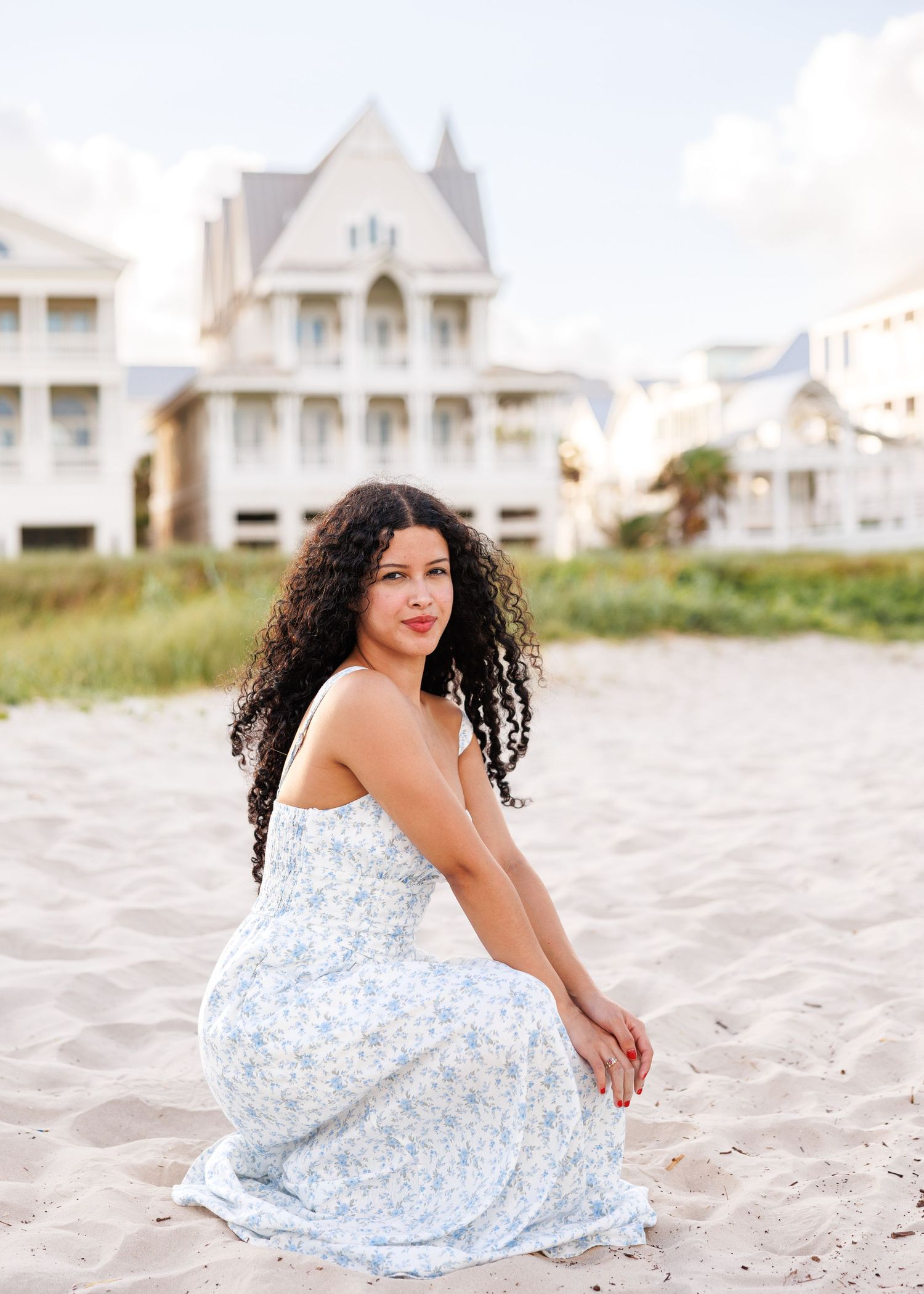 A woman in a patterned blue dress crouches gracefully on a sandy beach with white beachfront houses in the background.