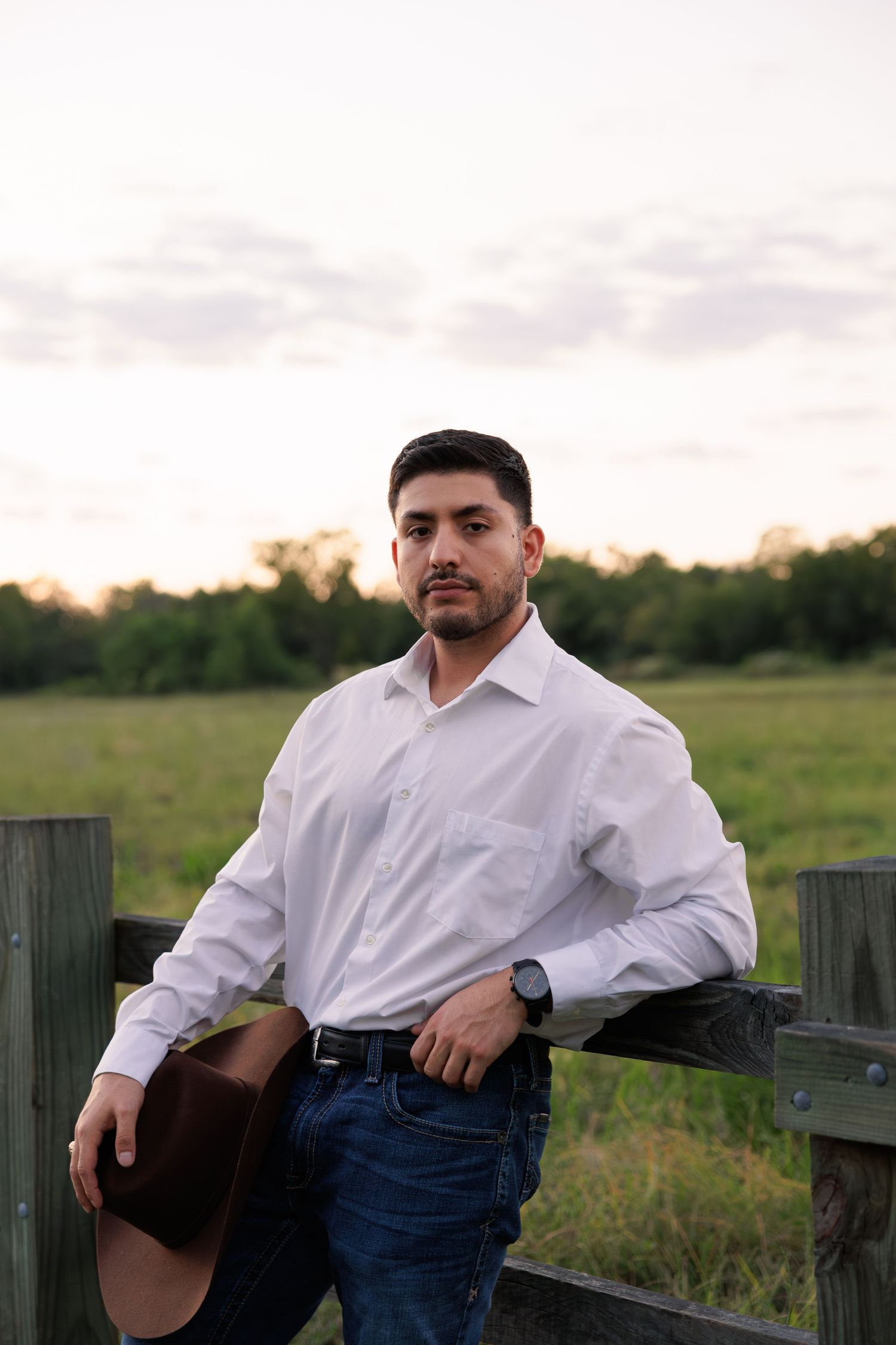 Man in white dress shirt and jeans leans against wooden fence in rural outdoor setting.