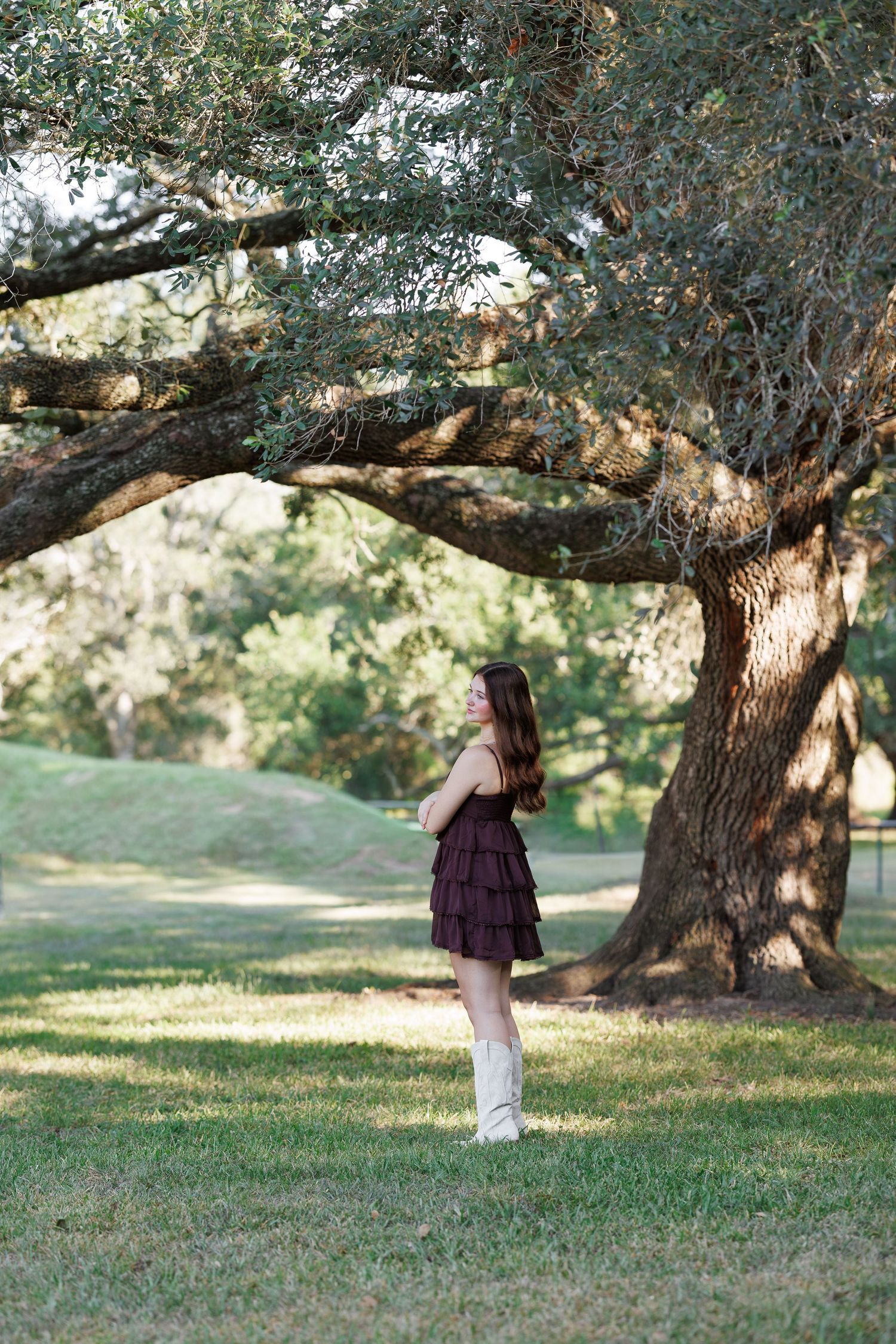 Young woman in a burgundy ruffled dress and white cowboy boots standing beneath a large live oak tree on a sunlit grassy lawn.