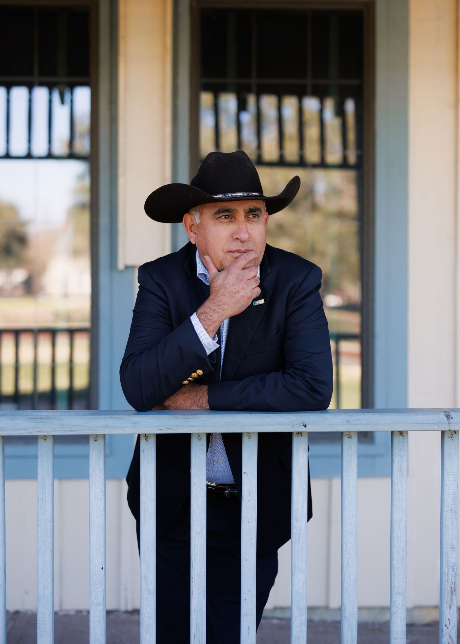 Person in a black cowboy hat and suit looking through white porch railings at ranch-style building exterior.