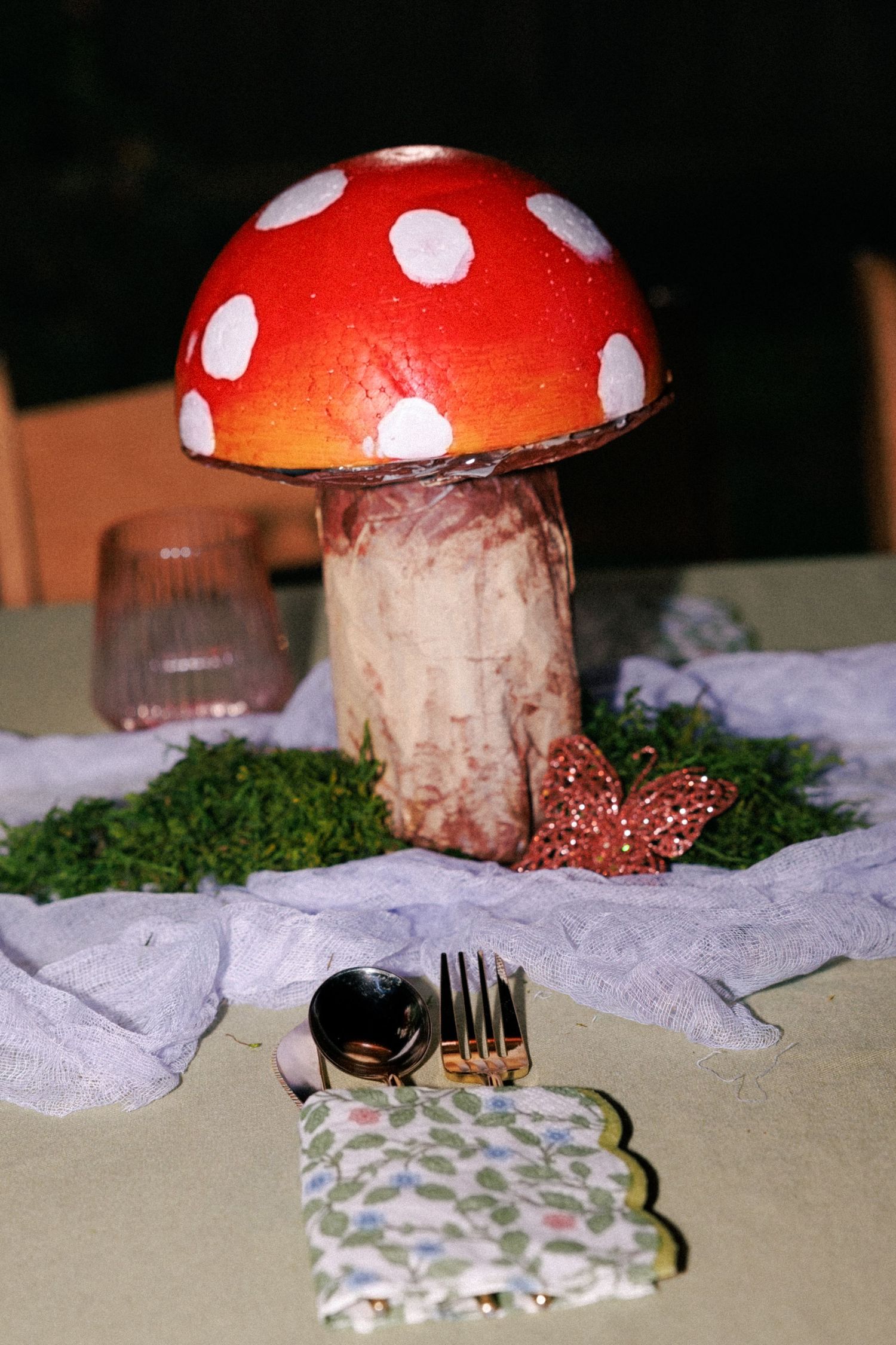 Red and white polka dot mushroom centerpiece on moss with gold fork, spoon, floral napkin, and pink glass at a themed event.