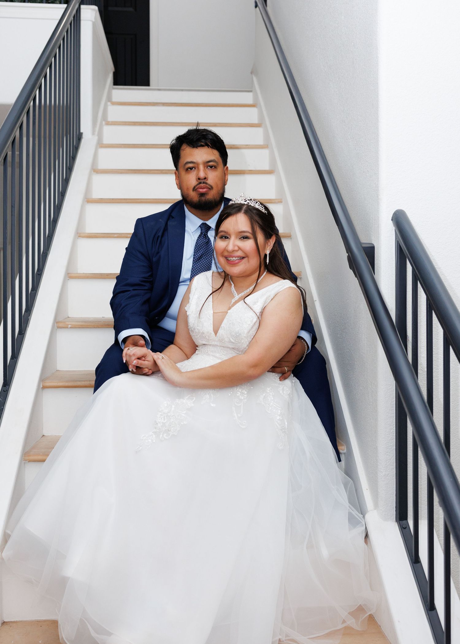 A couple in wedding attire sits together on white stairs with black railings.