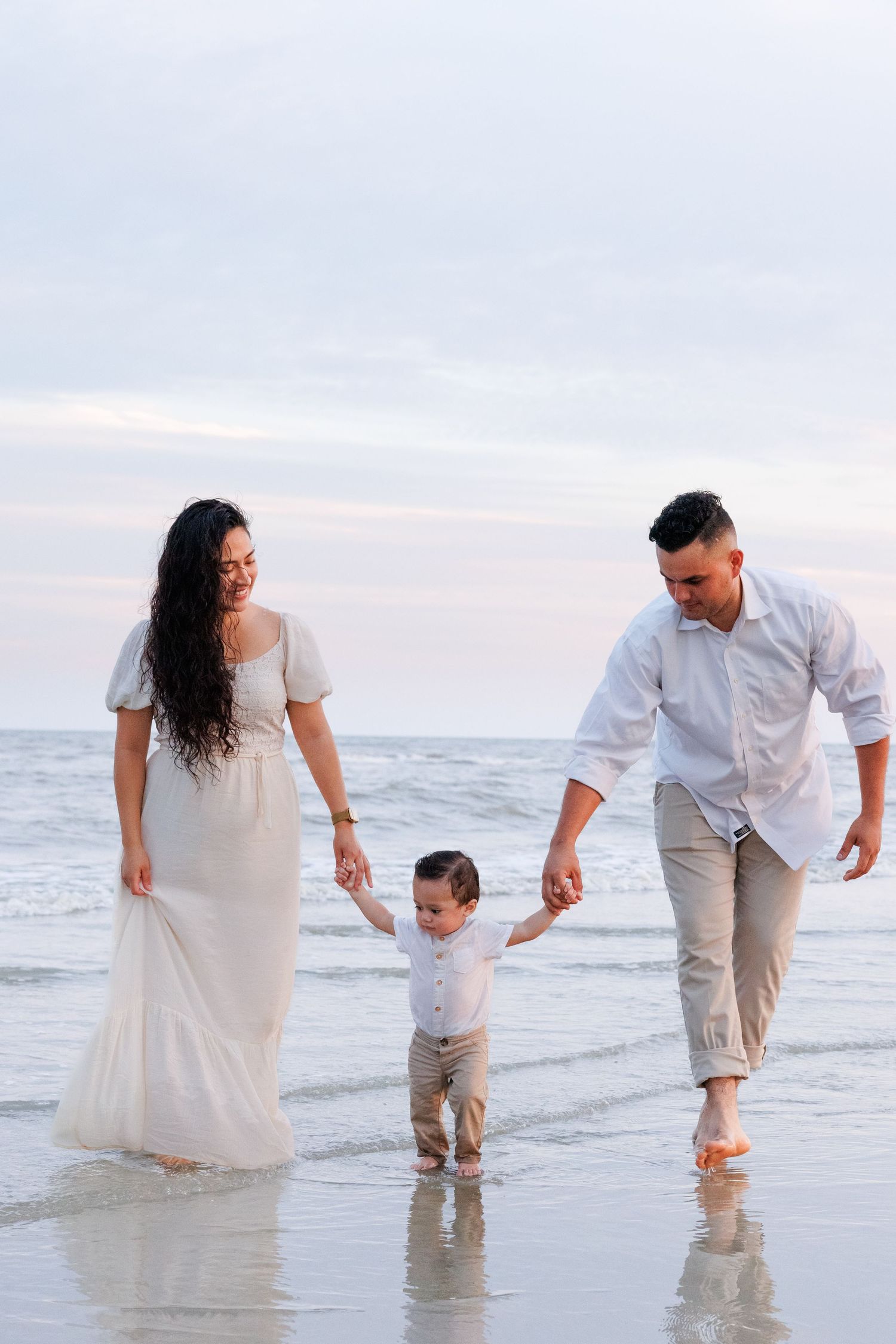 Family wearing white and beige outfits plays together at the beach during sunset while splashing in the ocean waves.