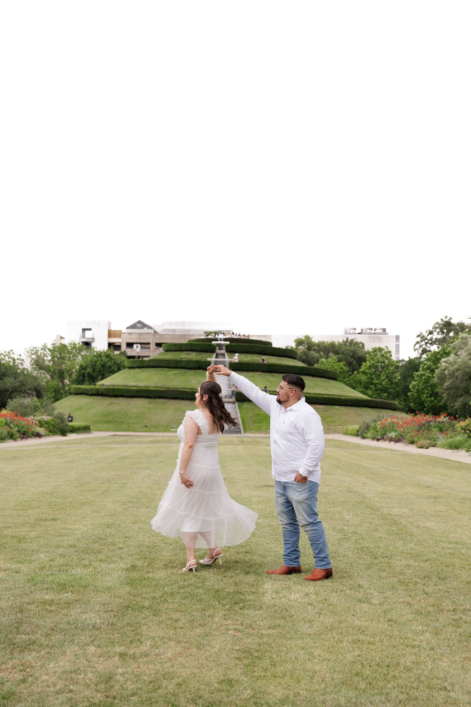 A romantic couple dancing together on a lush green lawn with landscaped gardens and buildings in the background.