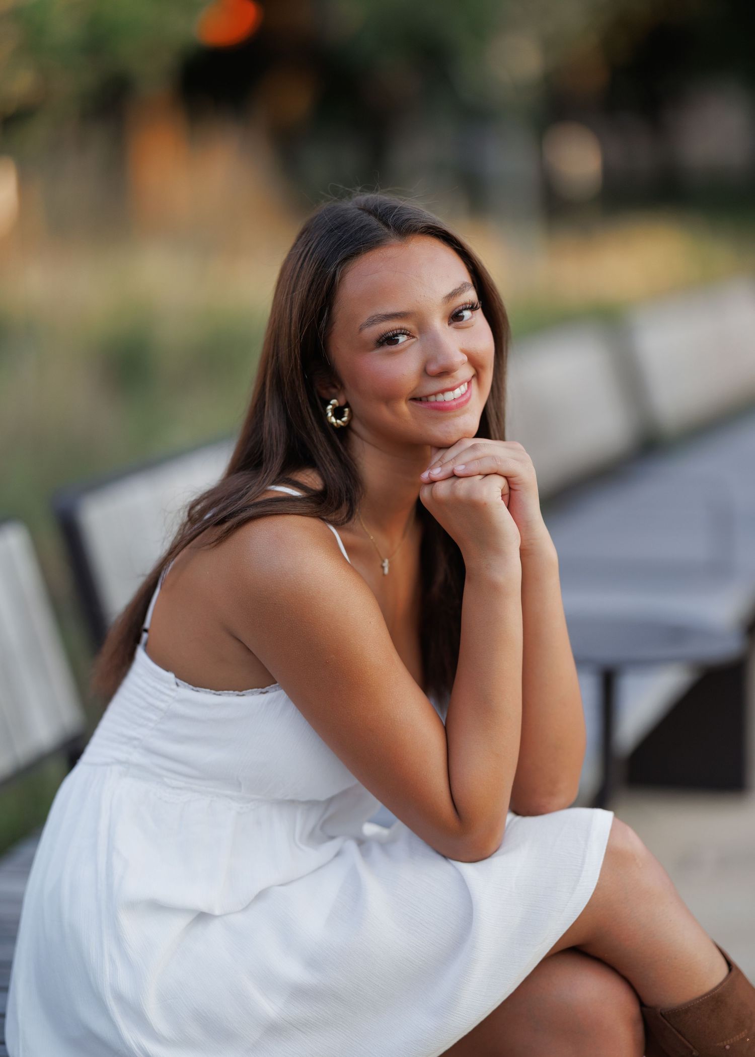 Smiling young woman with long brown hair, gold hoop earrings and a white sundress sits on an outdoor bench with her hands under her chin.