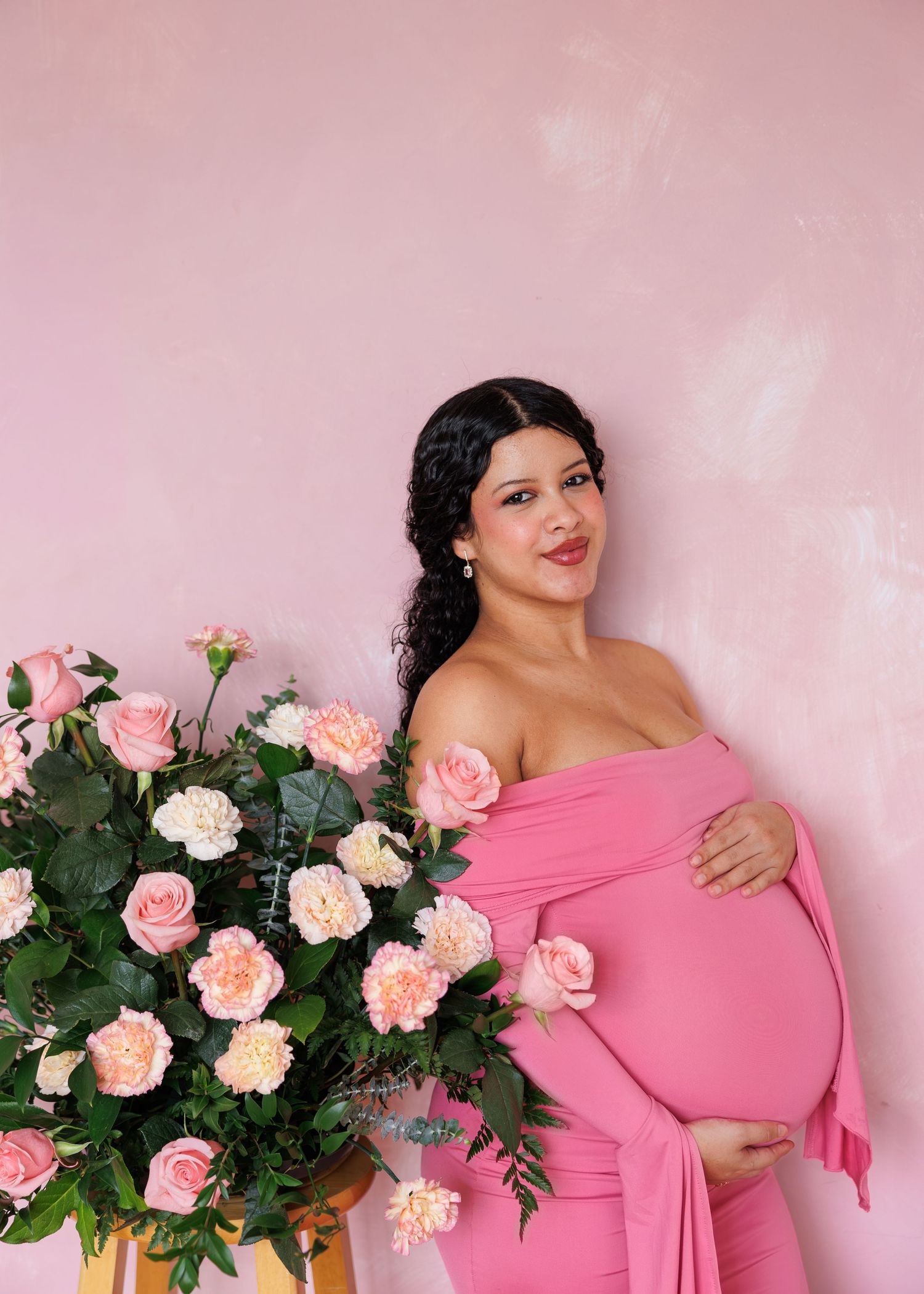 Pregnant woman in pink off-shoulder dress poses with pink and white roses against a soft pink wall.