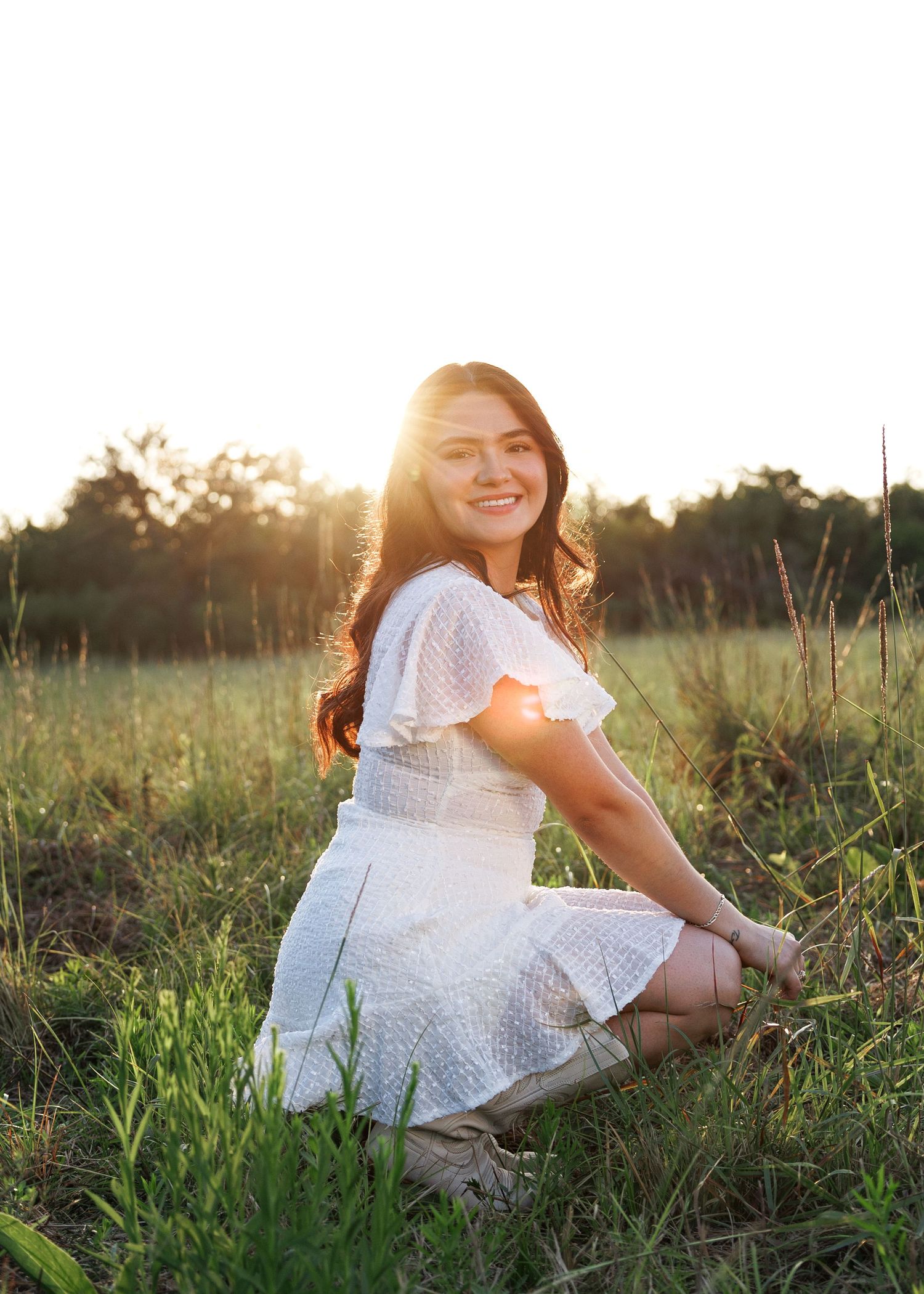 A person in a white eyelet dress sits gracefully in a sunlit meadow during golden hour.