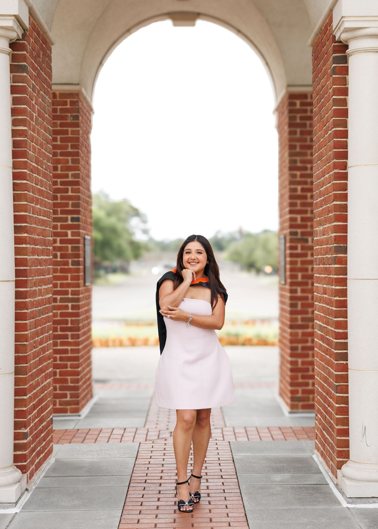 Young woman in white dress holding graduation cap smiles under brick archway on college campus.