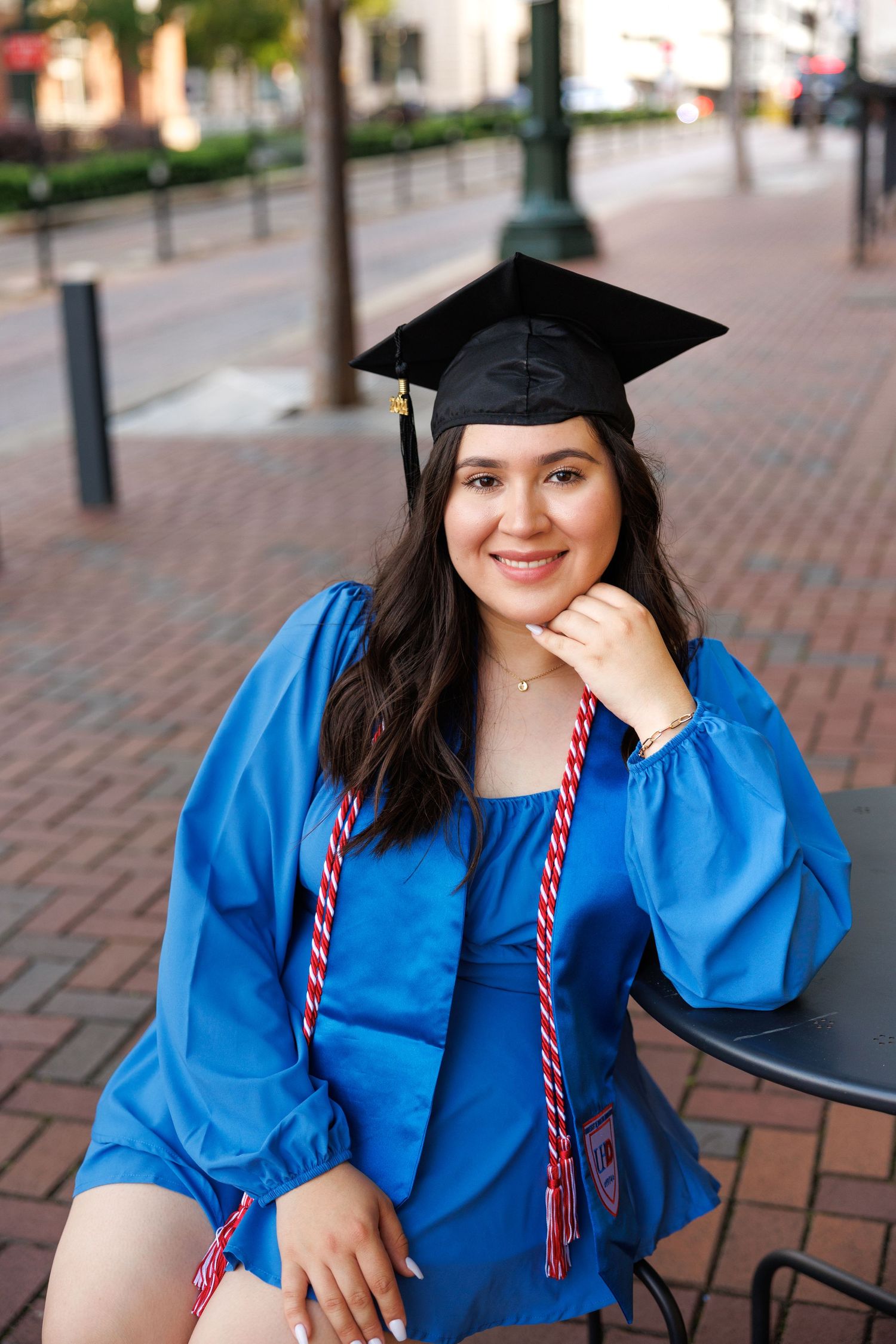 Graduate in bright blue commencement gown and mortarboard cap poses outdoors on brick sidewalk.